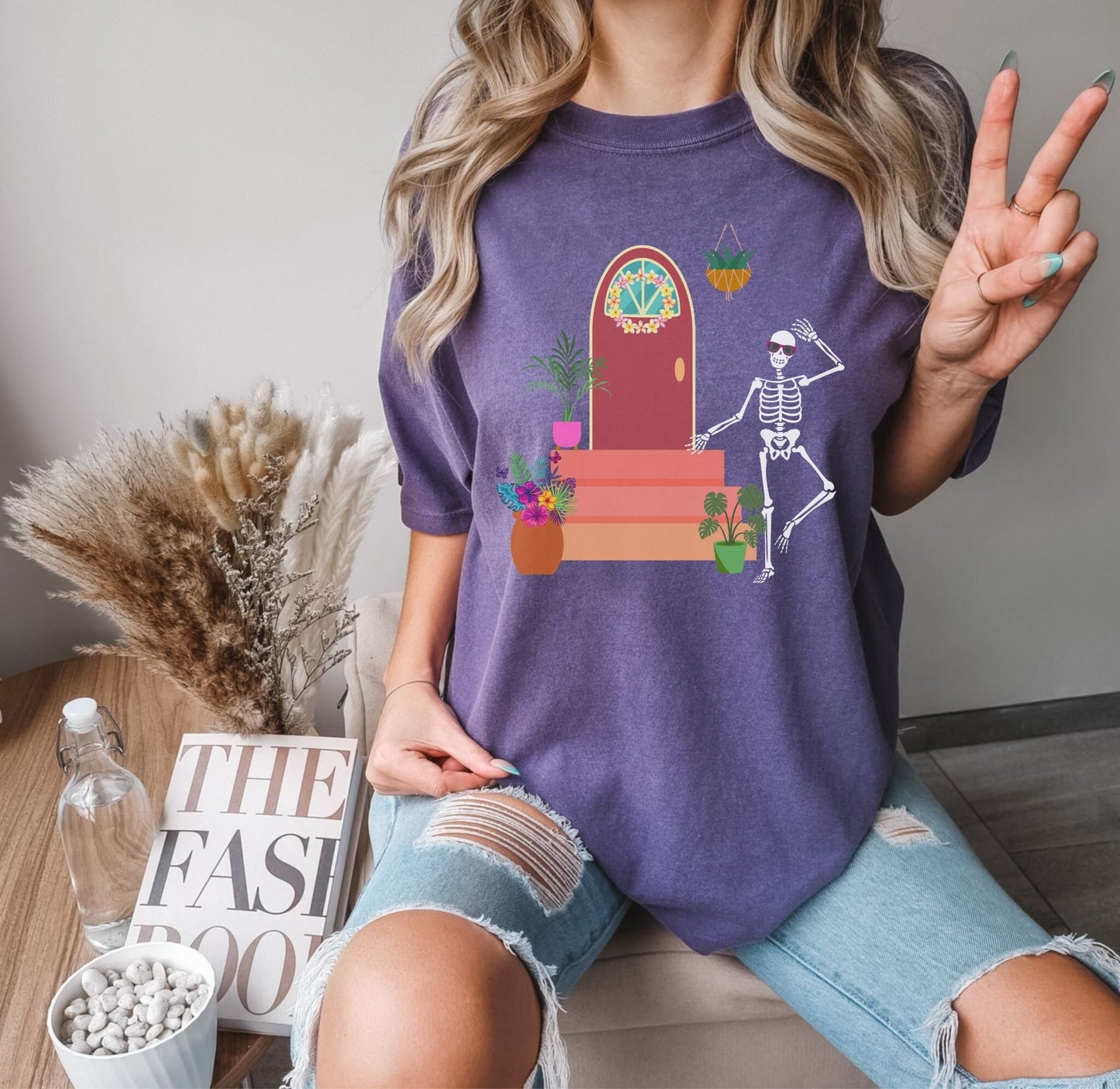 A woman wearing a cute, vintage midnight colored shirt with a skeleton wearing red sunglasses walking out the front door of their house. The front stoop has colorful plants and flowers and the front door has a pink and yellow flower wreath hanging.