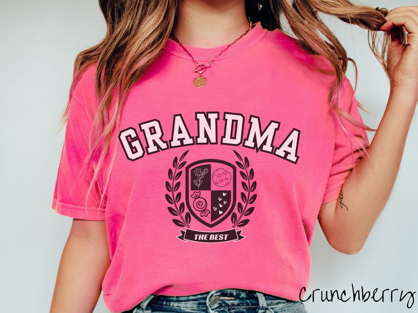 A woman wearing a cute crunchberry colored shirt with text saying Grandma across the front with a varsity logo underneath with the words The Best centered underneath the logo.