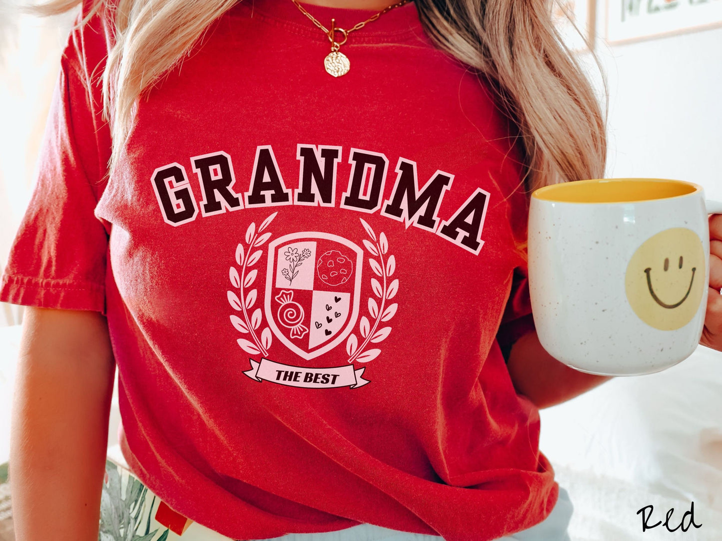 A woman wearing a cute red colored shirt with text saying Grandma across the front with a varsity logo underneath with the words The Best centered underneath the logo.