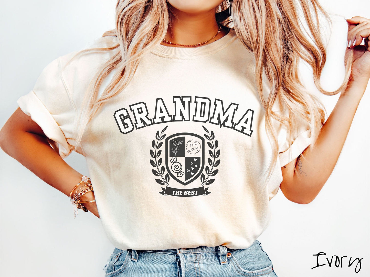 A woman wearing a cute ivory colored shirt with text saying Grandma across the front with a varsity logo underneath with the words The Best centered underneath the logo.