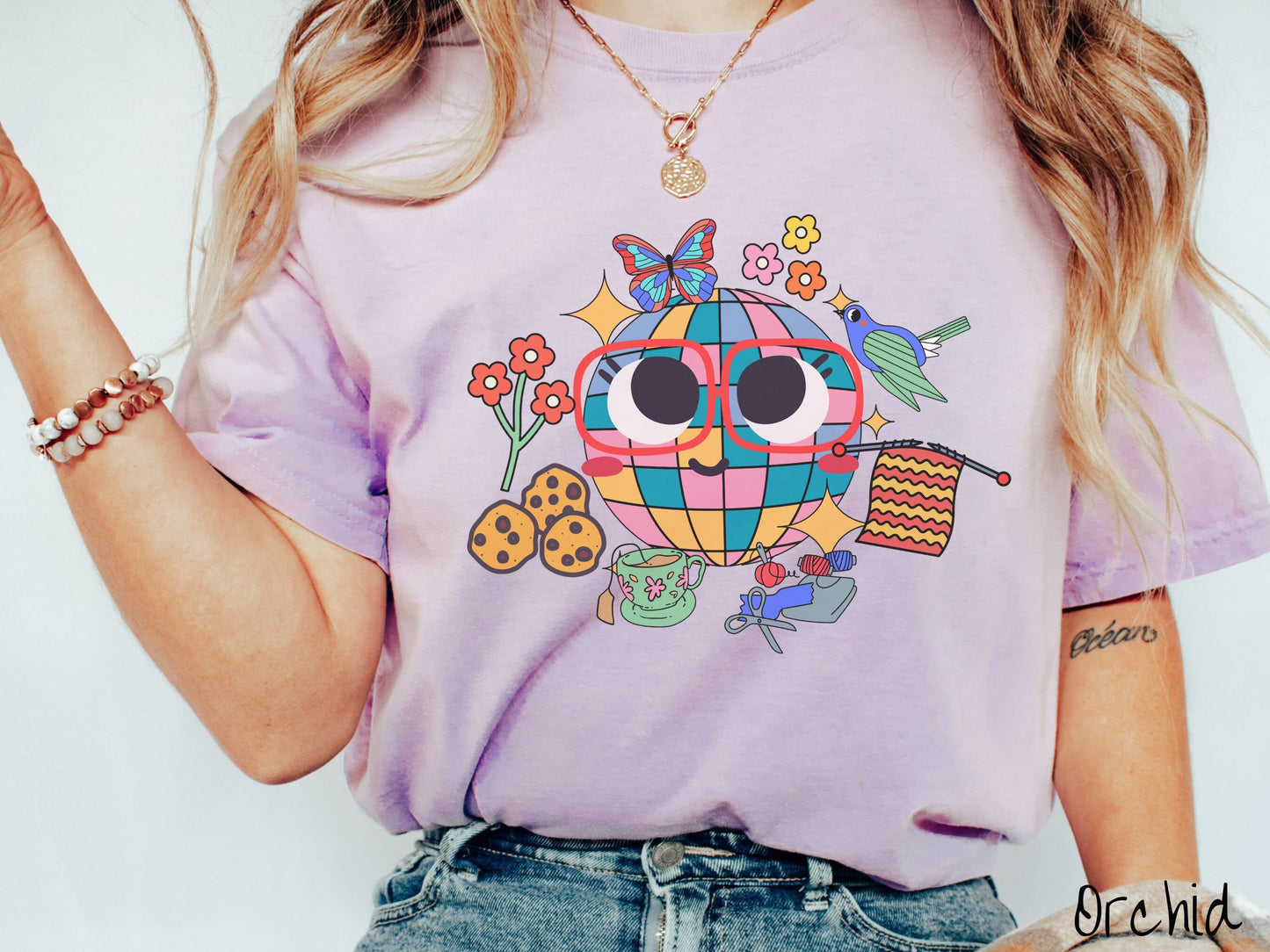 A woman wearing a cute orchid colored shirt with a smiling face and cookies, tea, flowers, butterflies, and birds mixed within each other.