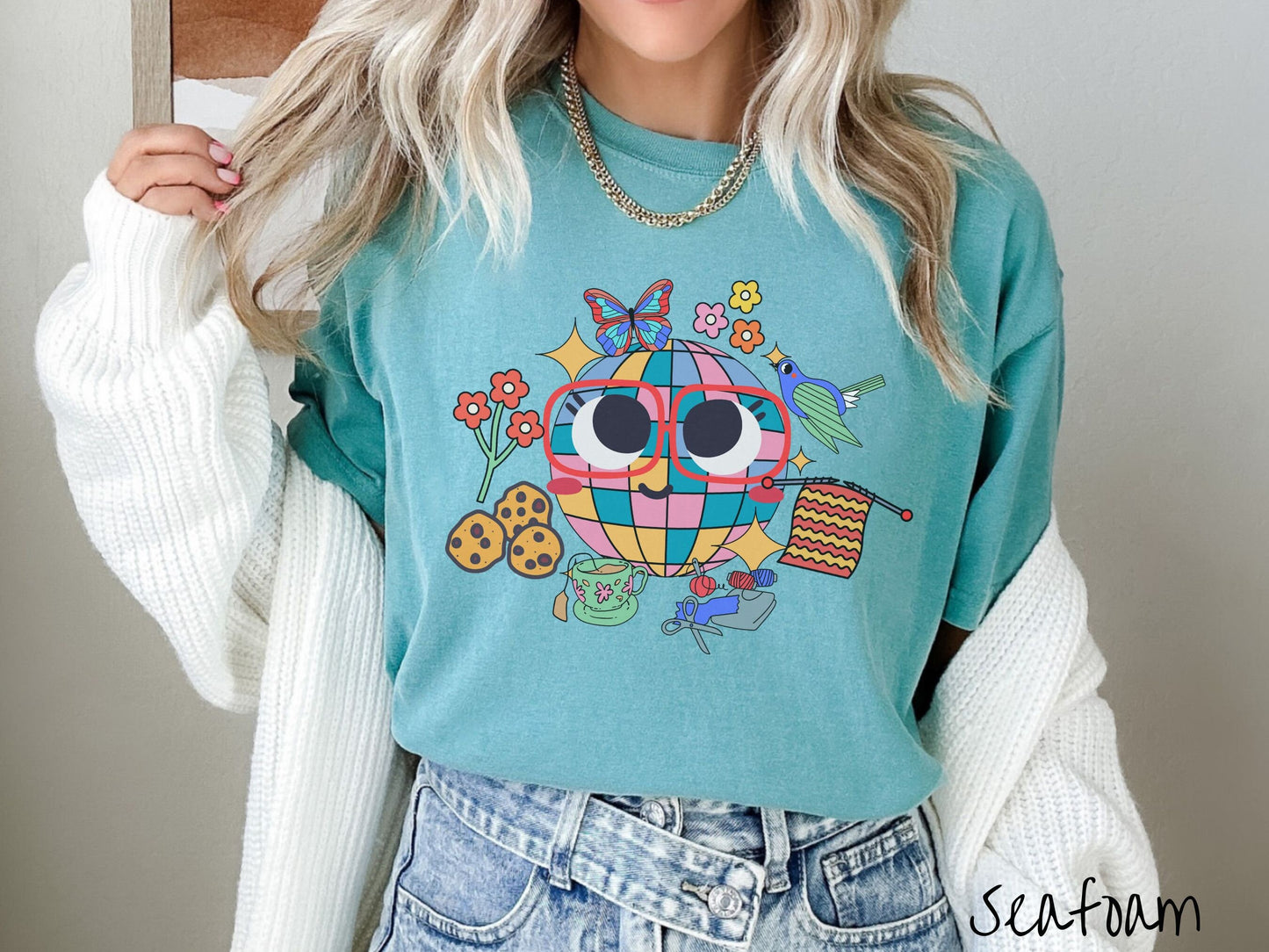 A woman wearing a cute seafoam colored shirt with a smiling face and cookies, tea, flowers, butterflies, and birds mixed within each other.