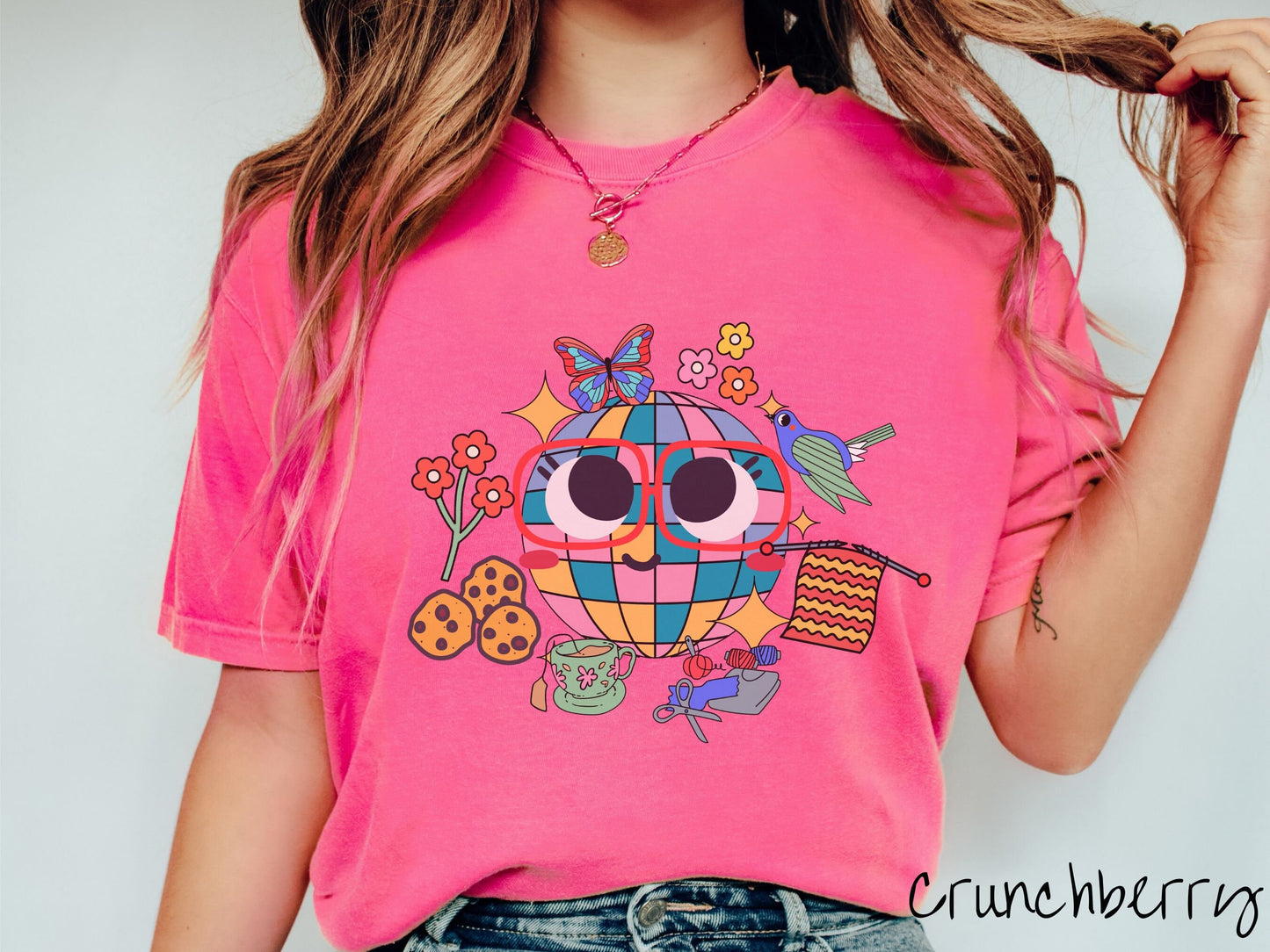 A woman wearing a cute crunchberry colored shirt with a smiling face and cookies, tea, flowers, butterflies, and birds mixed within each other.