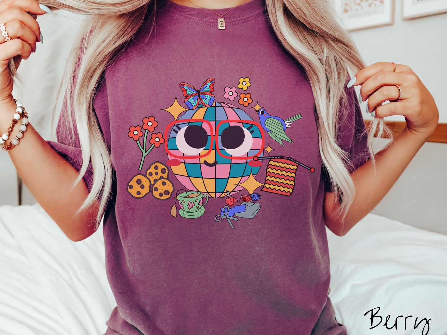 A woman wearing a cute berry colored shirt with a smiling face and cookies, tea, flowers, butterflies, and birds mixed within each other.