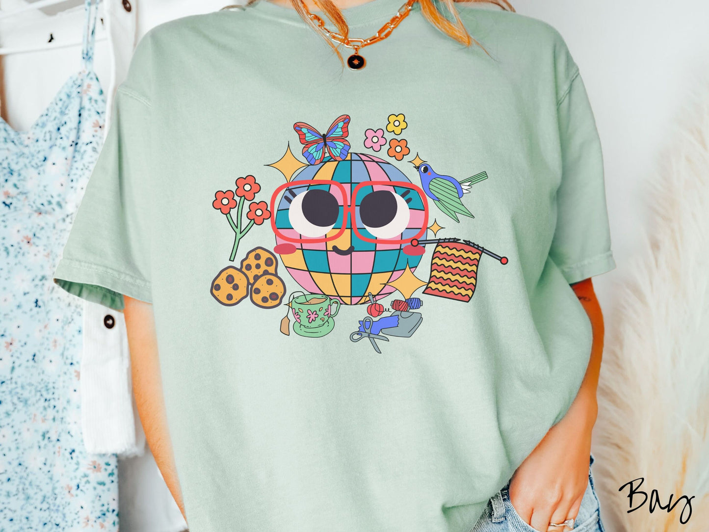 A woman wearing a cute bay colored shirt with a smiling face and cookies, tea, flowers, butterflies, and birds mixed within each other.