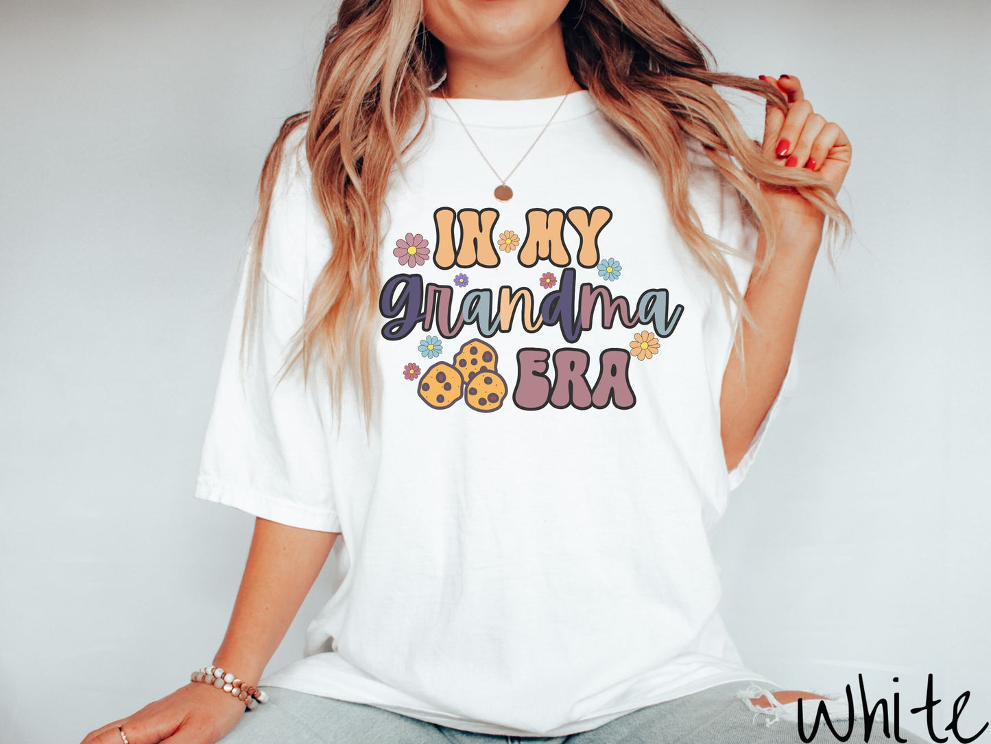 A woman wearing a cute white shirt with the text saying In My Grandma era with flowers and chocolate chip cookies mixed within the text.
