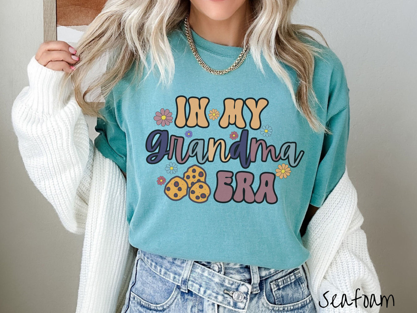 A woman wearing a cute cyan shirt with the text saying In My Grandma Era with flowers and chocolate chip cookies mixed within the text.