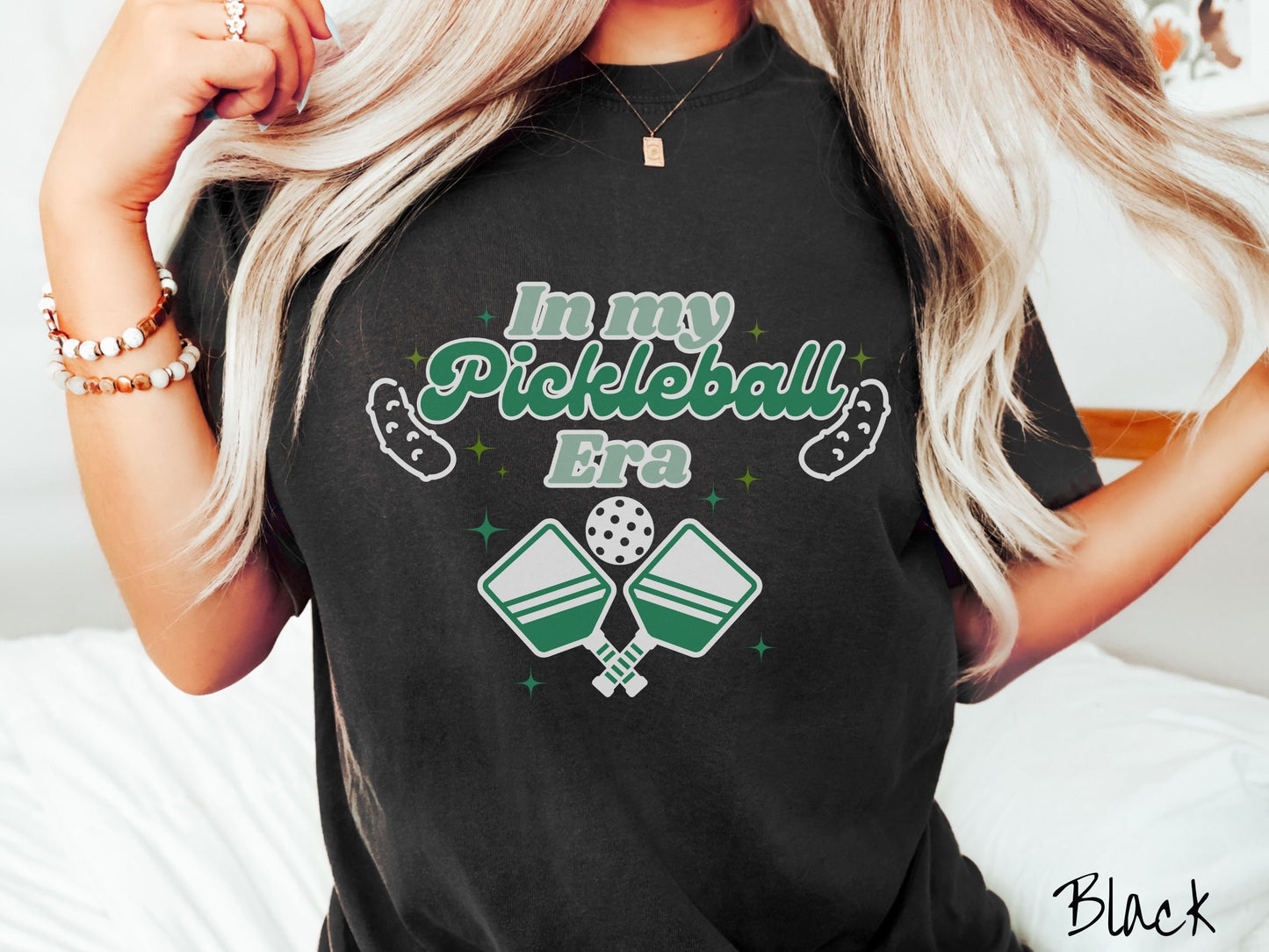 A woman wearing a cute black colored shirt with the text In My Pickleball Era across the top, and below are 2 pickleball paddles and a ball. There are pickles at the bottom left and right corners of the text.