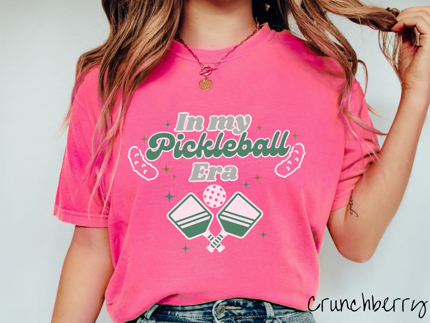 A woman wearing a cute crunchberry colored shirt with the text In My Pickleball Era across the top, and below are 2 pickleball paddles and a ball. There are pickles at the bottom left and right corners of the text.