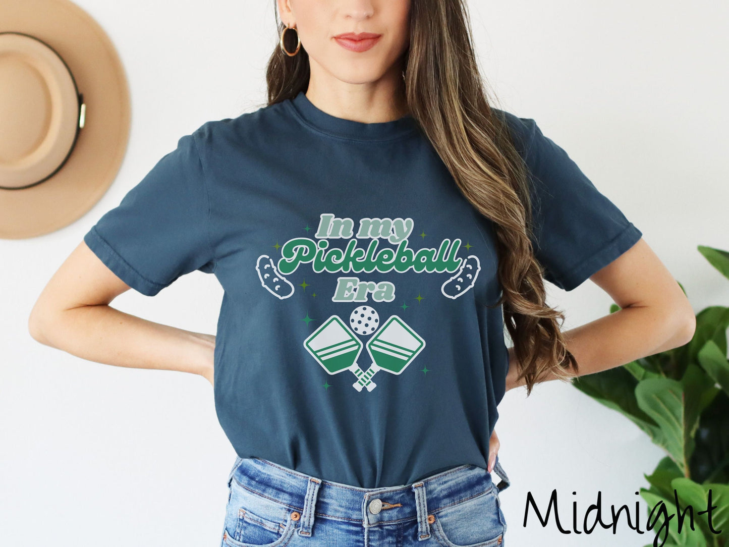 A woman wearing a cute midnight colored shirt with the text In My Pickleball Era across the top, and below are 2 pickleball paddles and a ball. There are pickles at the bottom left and right corners of the text.
