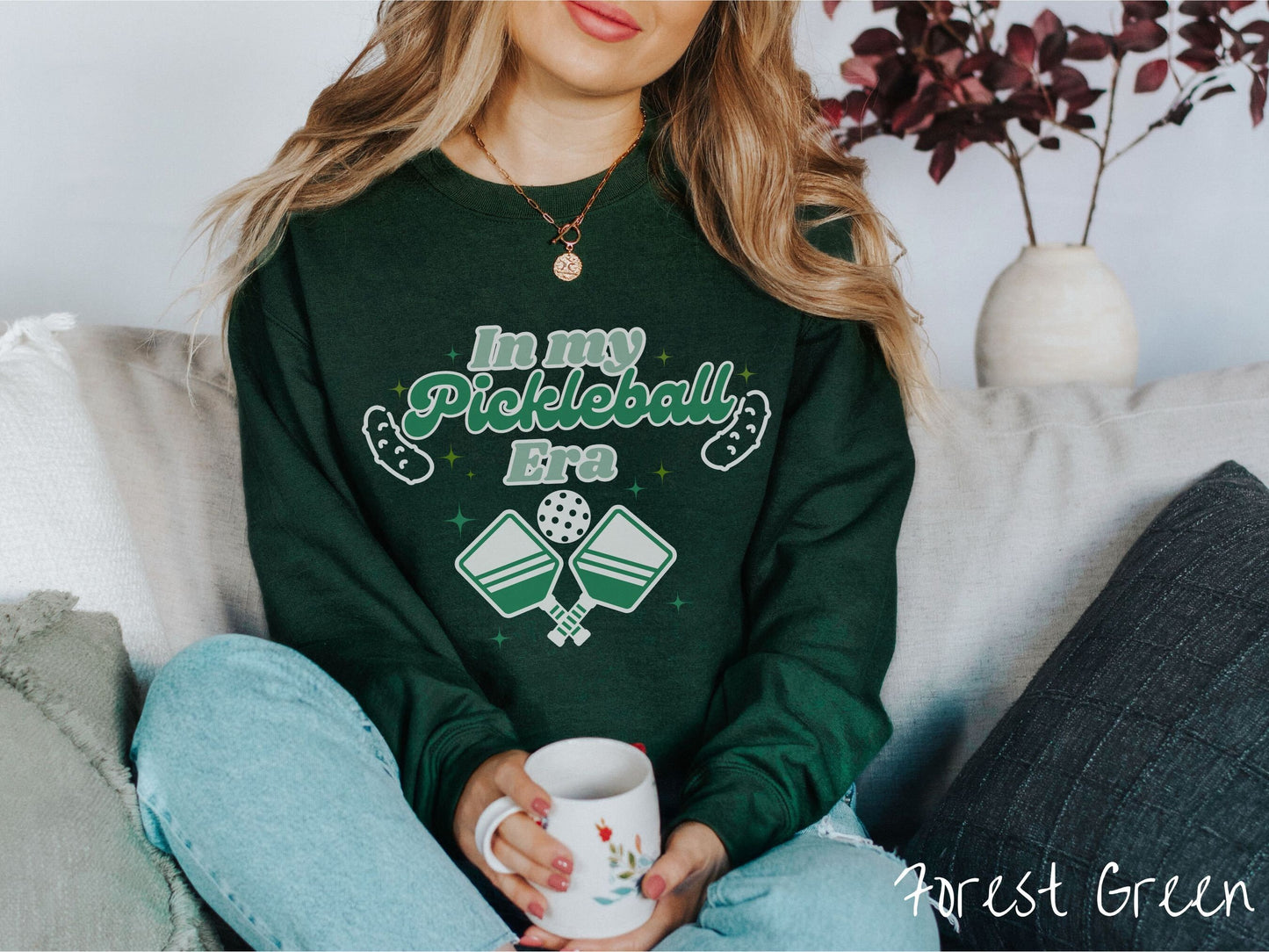 A woman wearing a cute forest green colored sweatshirt with the text In My Pickleball Era across the top, and below are 2 pickleball paddles and a ball. There are pickles at the bottom left and right corners of the text.