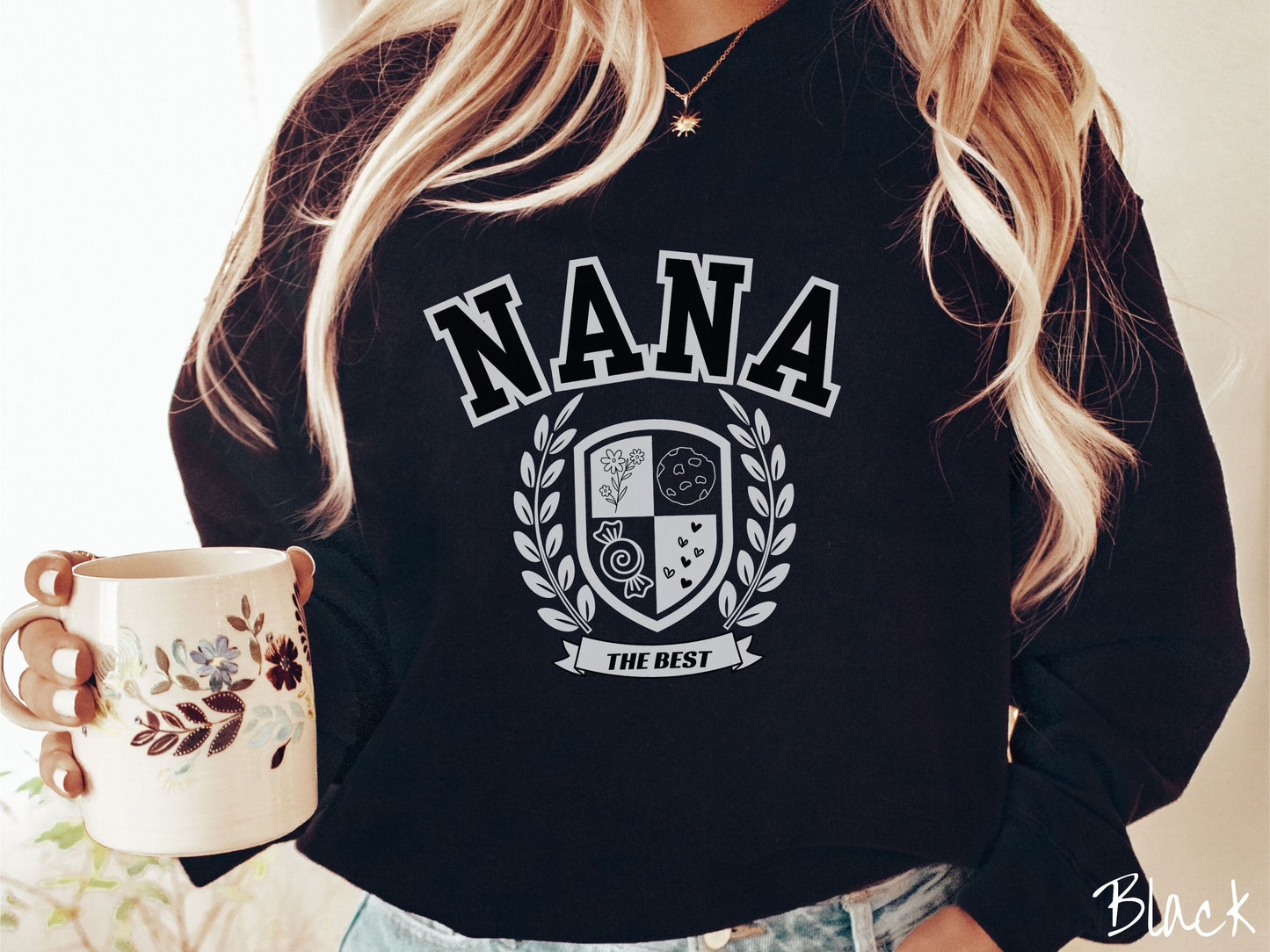 A woman wearing a cute black colored sweatshirt with text saying Nana across the front with a varsity logo underneath with the words The Best centered underneath the logo.