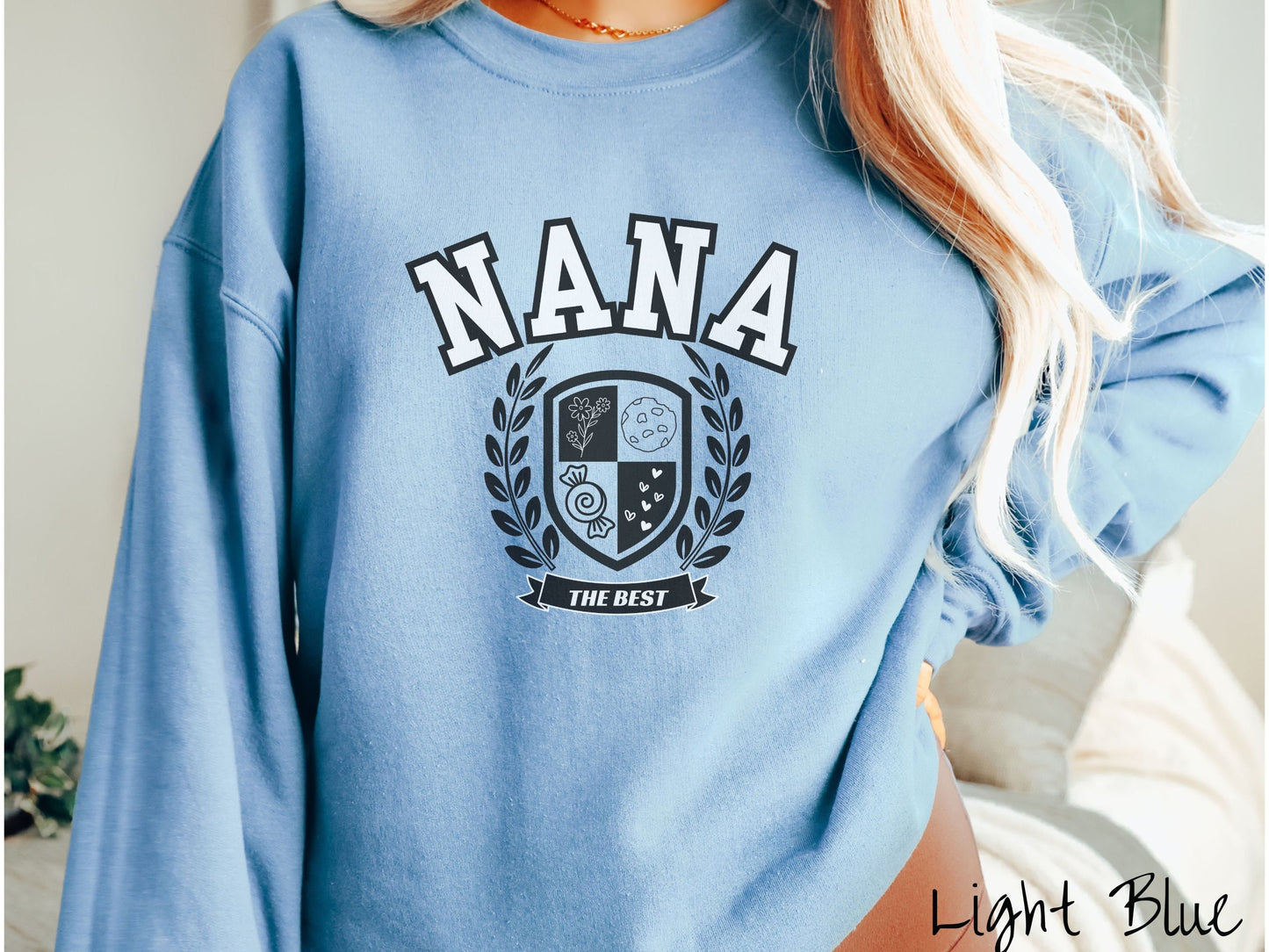 A woman wearing a cute light blue colored sweatshirt with text saying Nana across the front with a varsity logo underneath with the words The Best centered underneath the logo.