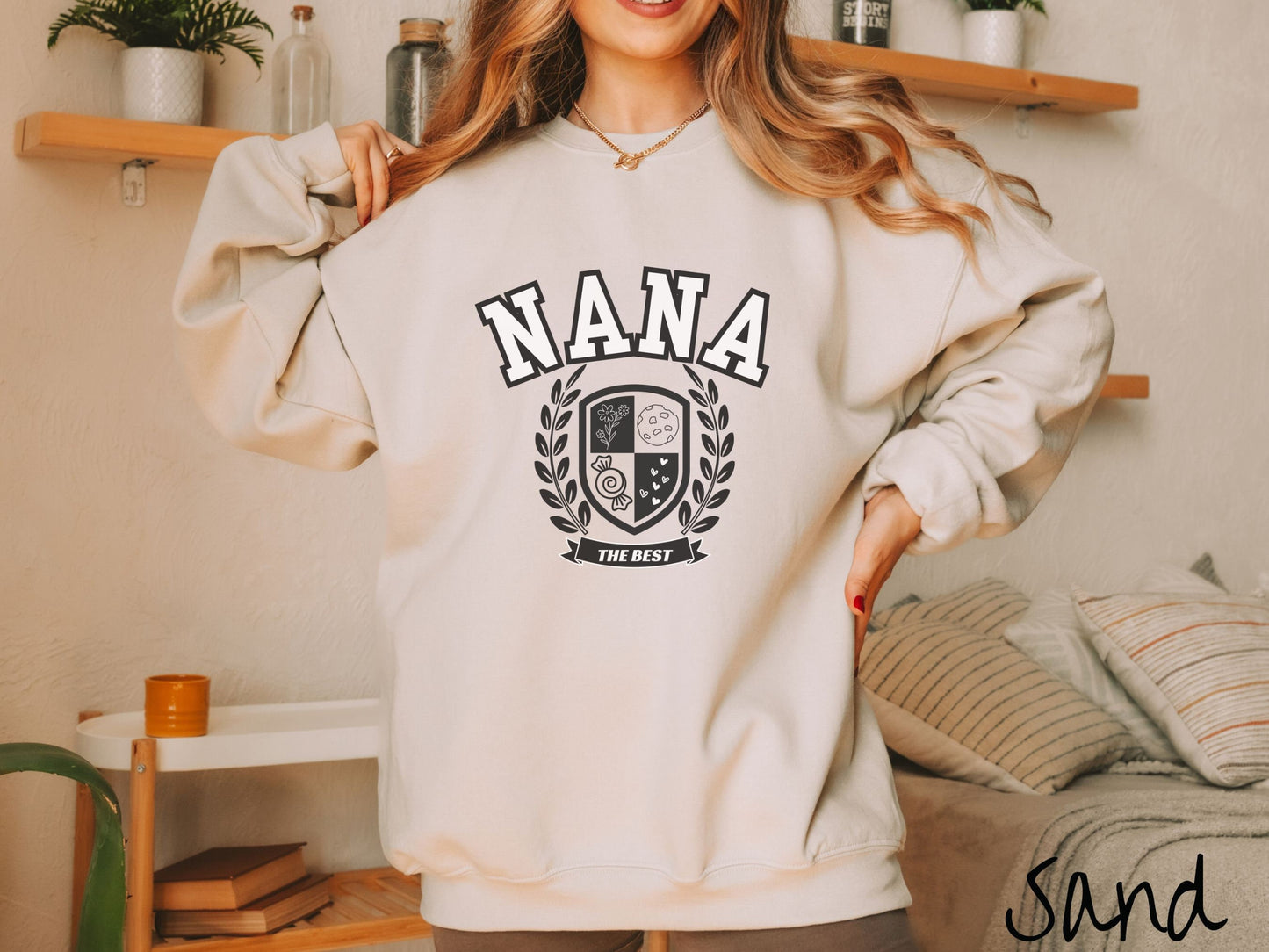 A woman wearing a cute sand colored sweatshirt with text saying Nana across the front with a varsity logo underneath with the words The Best centered underneath the logo.