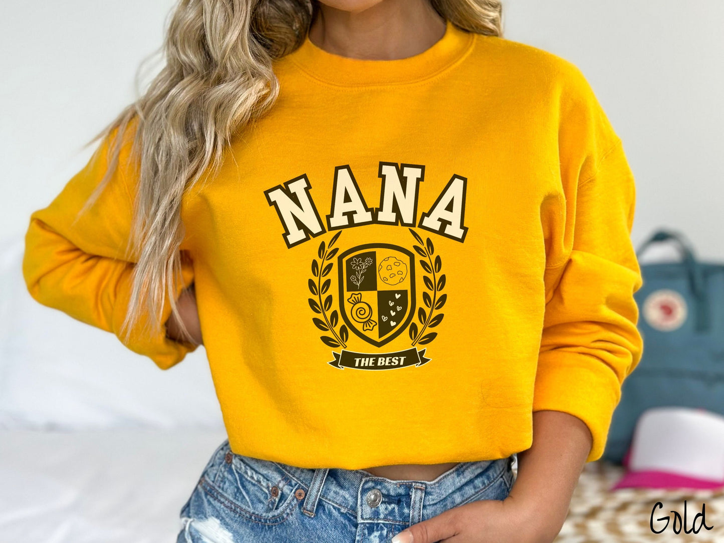 A woman wearing a cute gold colored sweatshirt with text saying Nana across the front with a varsity logo underneath with the words The Best centered underneath the logo.