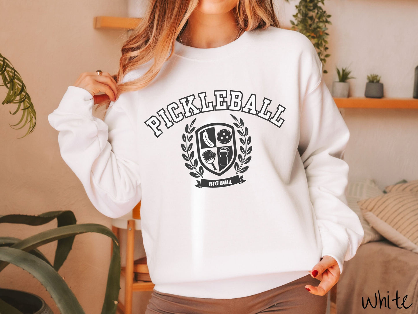 A woman wearing a cute white colored sweatshirt with the text Pickleball across the top, a varsity logo centered underneath, and the words Big Dill underneath the logo.