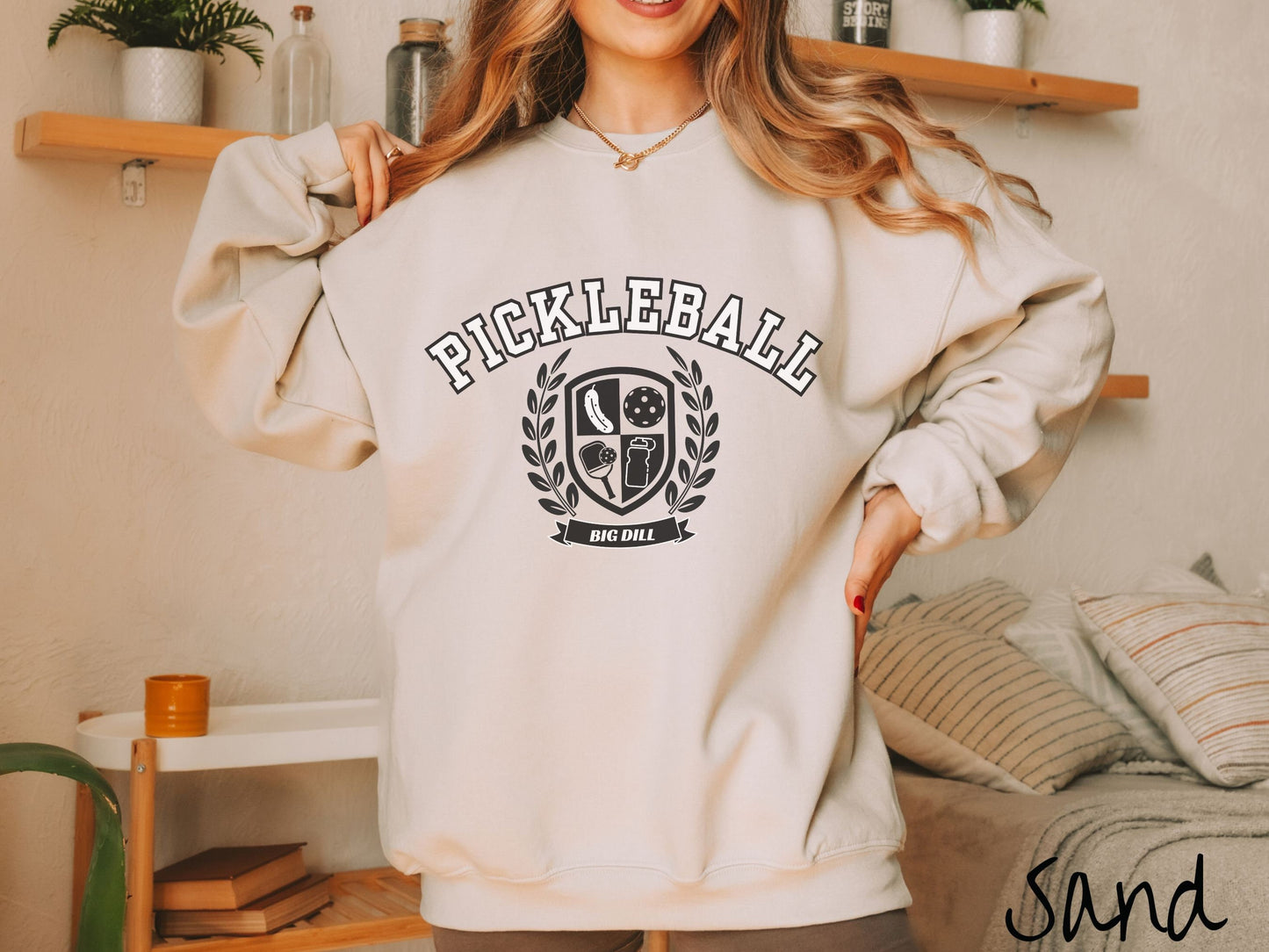 A woman wearing a cute sand colored sweatshirt with the text Pickleball across the top, a varsity logo centered underneath, and the words Big Dill underneath the logo.