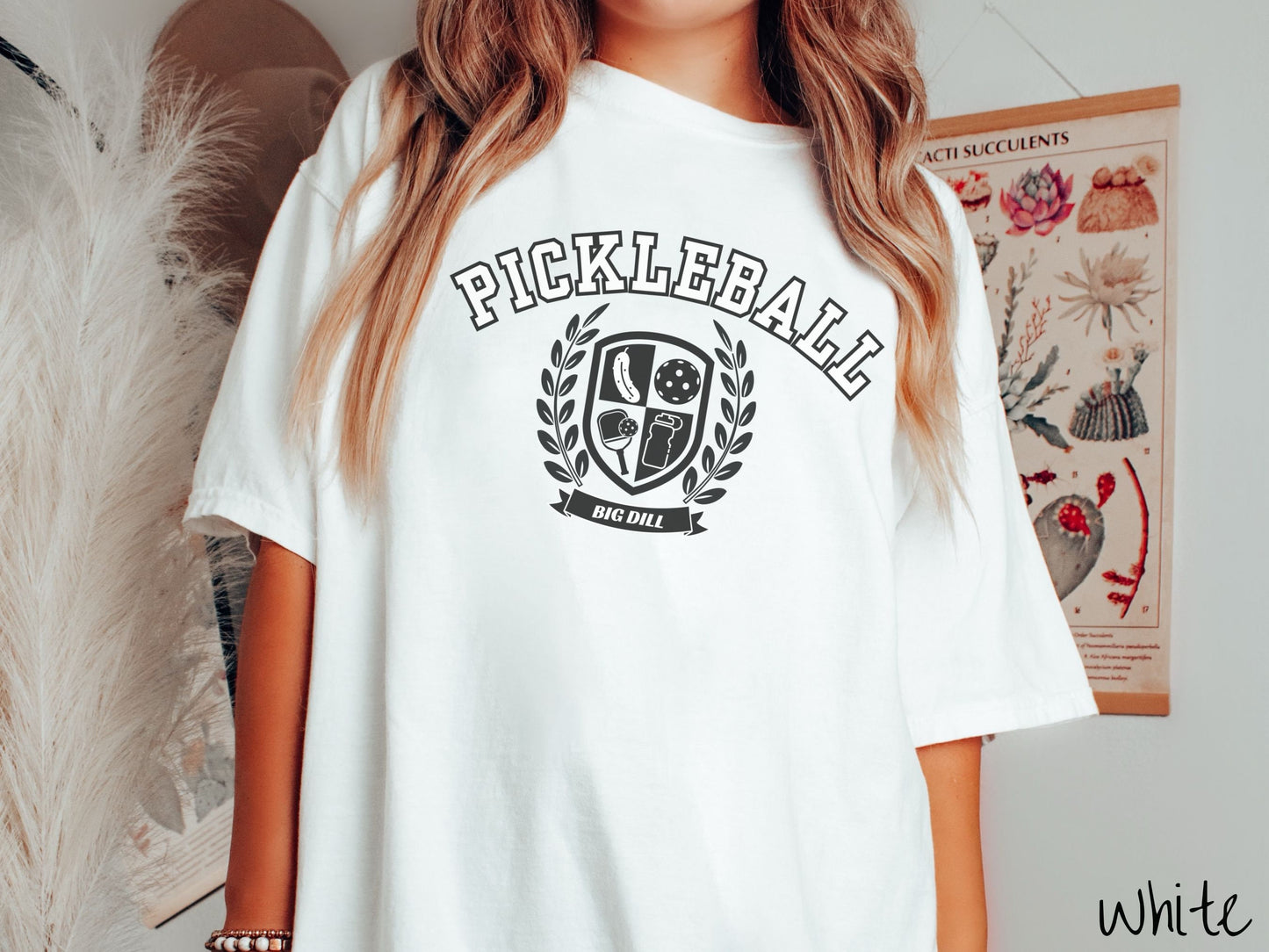 A woman wearing a cute white colored shirt with the text Pickleball across the top, a varsity logo centered underneath, and the words Big Dill underneath the logo.