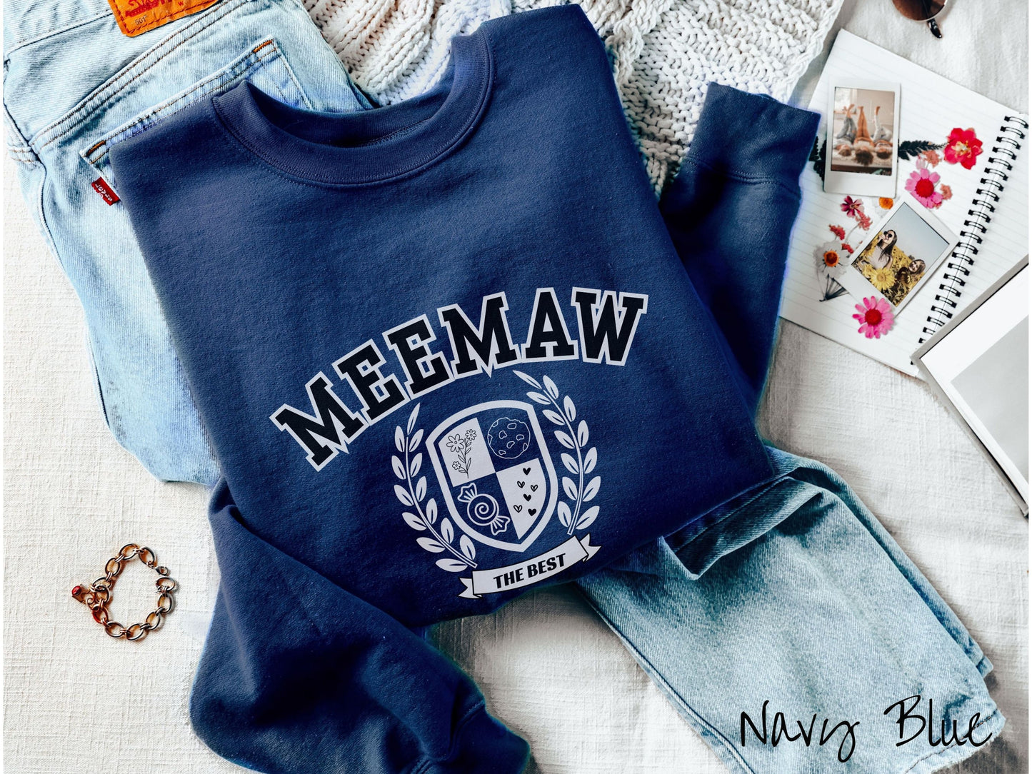 A woman wearing a cute navy blue colored sweatshirt with text saying Meemaw across the front with a varsity logo underneath with the words The Best centered underneath the logo.