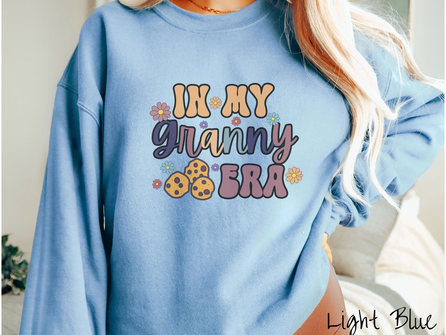 A woman wearing a cute light blue colored sweatshirt with text on the front saying In My Granny Era, with flowers and chocolate chip cookies mixed within the text.