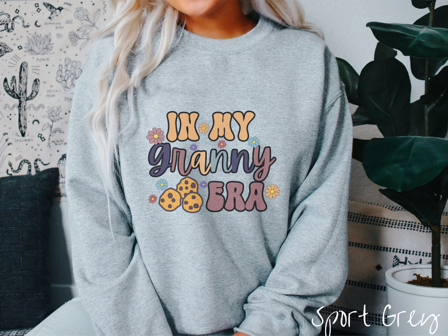 A woman wearing a cute sport grey colored sweatshirt with text on the front saying In My Granny Era, with flowers and chocolate chip cookies mixed within the text.