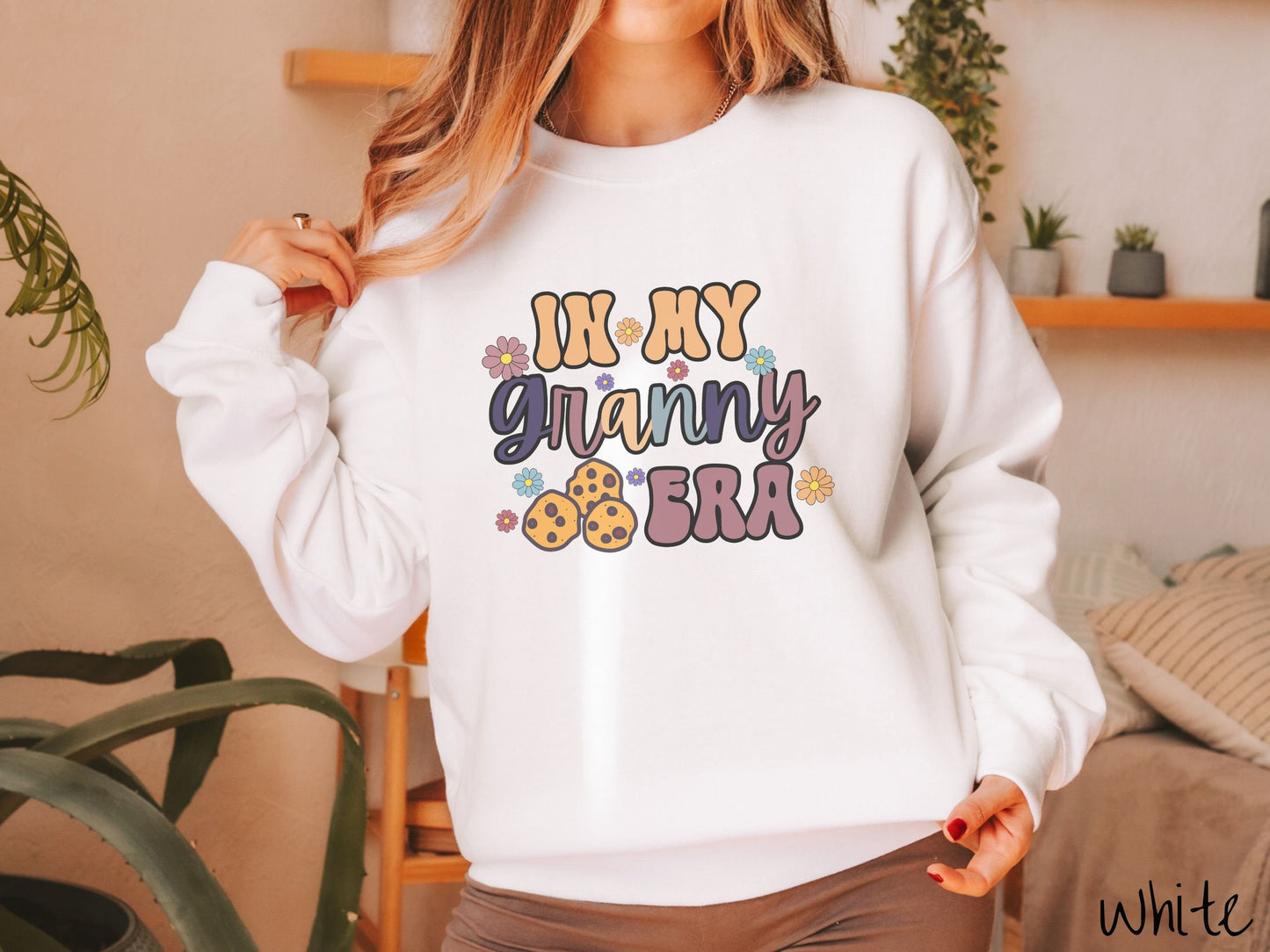 A woman wearing a cute white colored sweatshirt with text on the front saying In My Granny Era, with flowers and chocolate chip cookies mixed within the text.