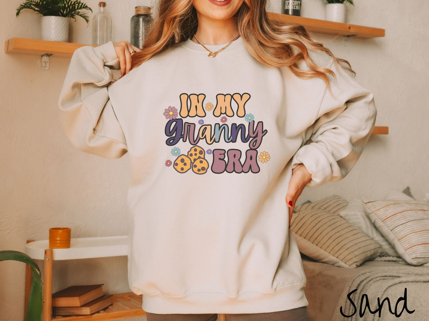 A woman wearing a cute sand colored sweatshirt with text on the front saying In My Granny Era, with flowers and chocolate chip cookies mixed within the text.