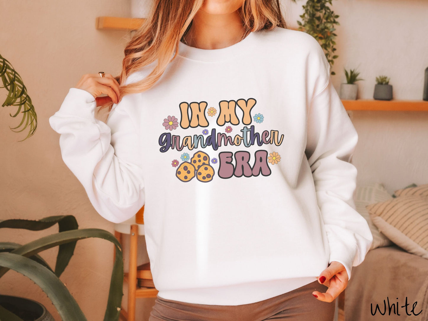 A woman wearing a cute white colored sweatshirt with text on the front saying In My Grandmother Era, with flowers and chocolate chip cookies mixed within the text.