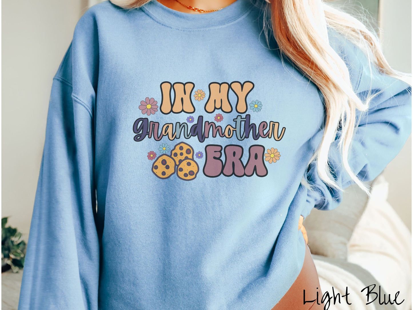 A woman wearing a cute light blue colored sweatshirt with text on the front saying In My Grandmother Era, with flowers and chocolate chip cookies mixed within the text.
