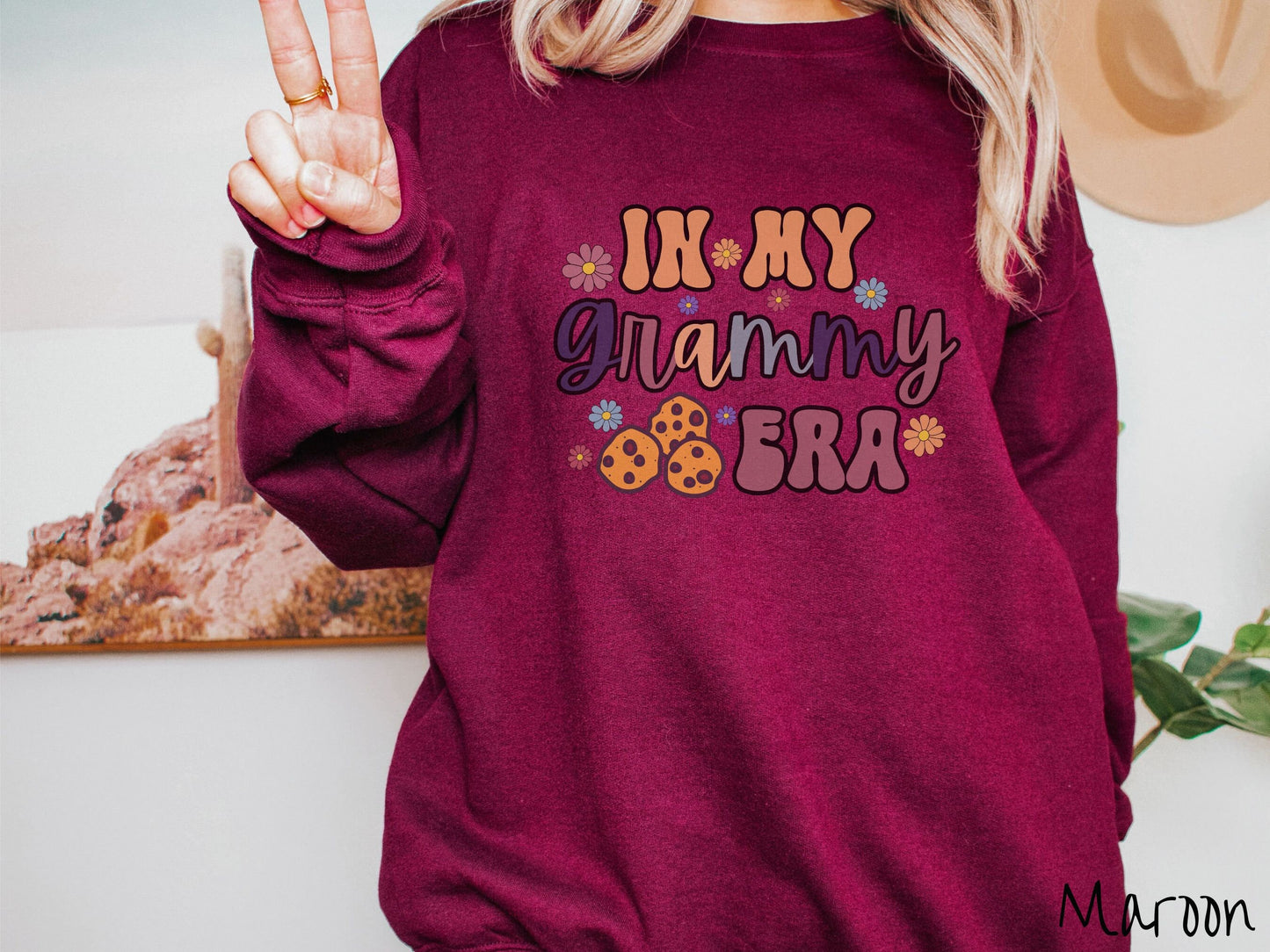 A woman wearing a cute, maroon sweatshirt with the text saying In My Grammy era with flowers and chocolate chip cookies mixed within the text.