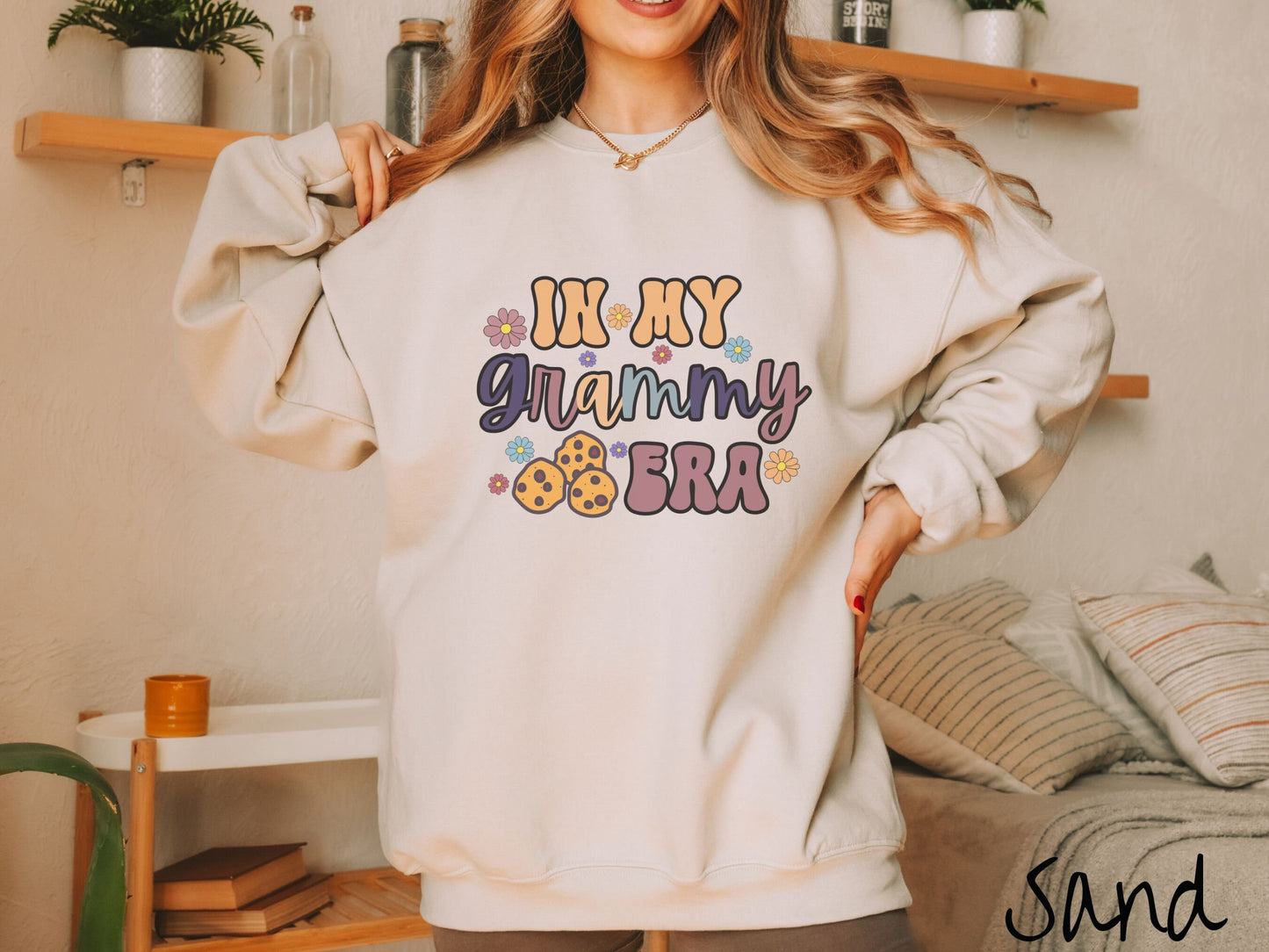 A woman wearing a cute, cream sweatshirt with the text saying In My Grammy era with flowers and chocolate chip cookies mixed within the text.