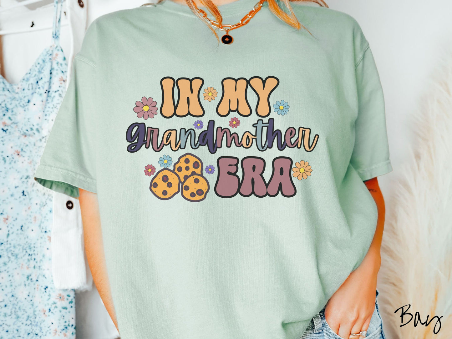 A woman wearing a cute bay colored shirt with text on the front saying In My Grandmother Era, with flowers and chocolate chip cookies mixed within the text.