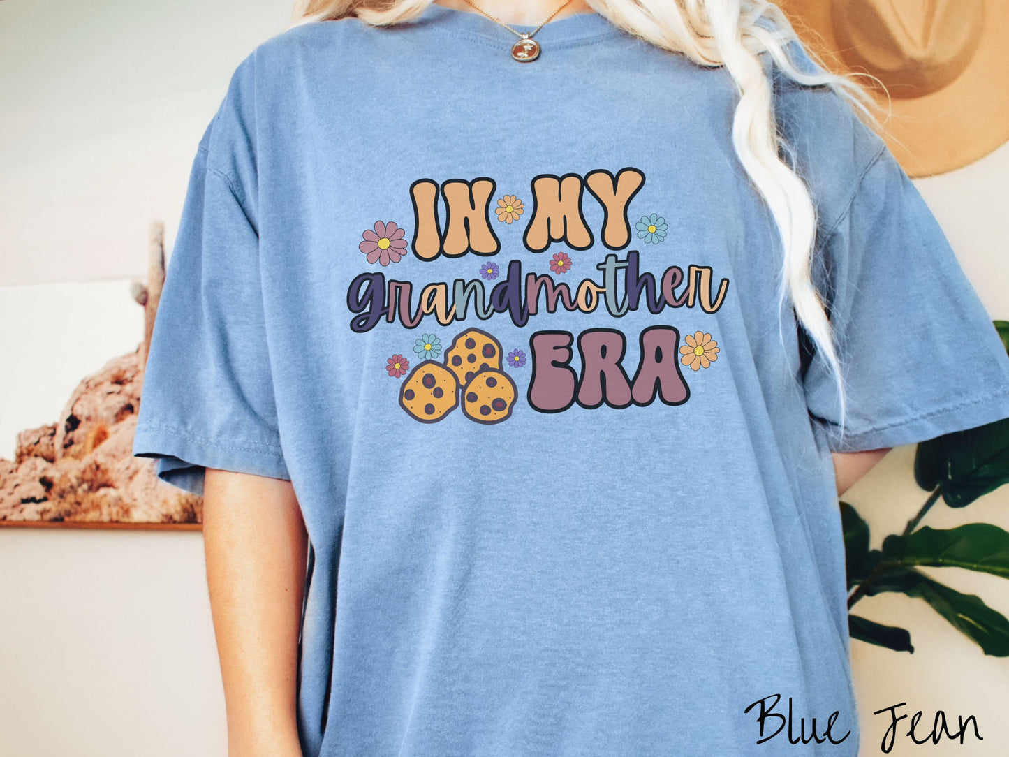 A woman wearing a cute blue jean colored shirt with text on the front saying In My Grandmother Era, with flowers and chocolate chip cookies mixed within the text.