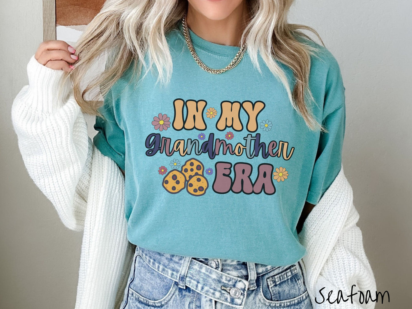 A woman wearing a cute seafoam colored shirt with text on the front saying In My Grandmother Era, with flowers and chocolate chip cookies mixed within the text.