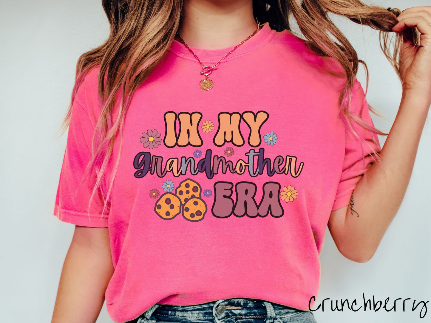 A woman wearing a crunchberry pink colored shirt with text on the front saying In My Grandmother Era, with flowers and chocolate chip cookies mixed within the text.