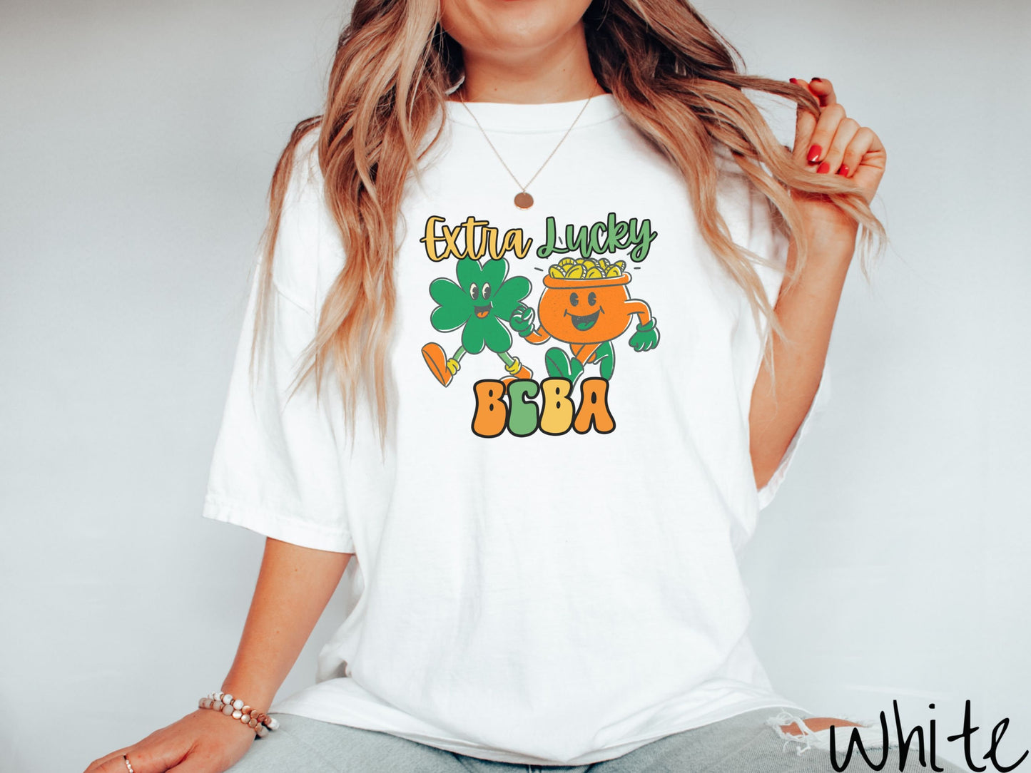 A woman wearing a vintage, white colored shirt with the text Extra Lucky BCBA in yellow, orange, and green font. Between the text are a green clover and orange pot of gold smiling and walking together in step.