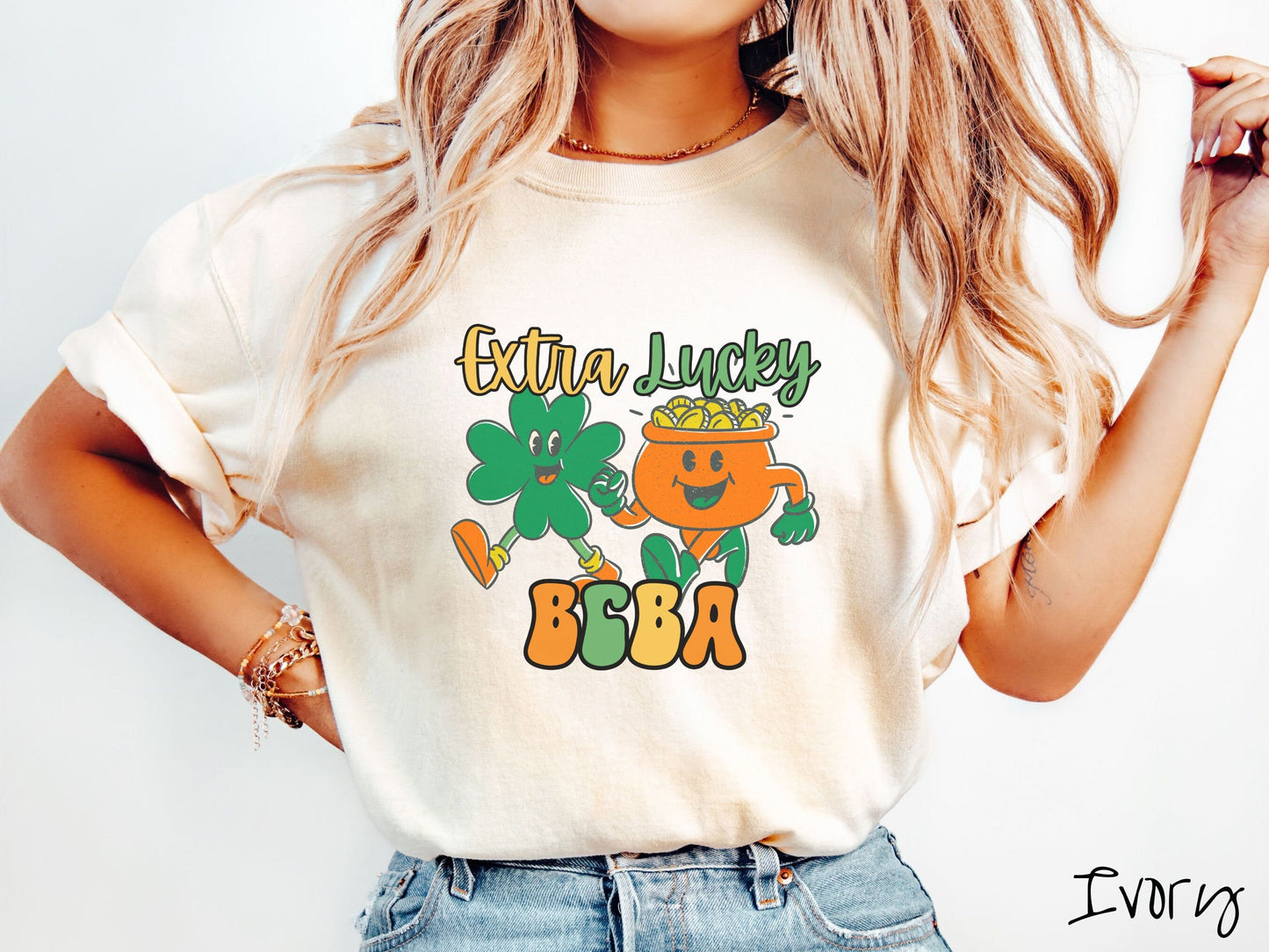 A woman wearing a vintage, ivory colored shirt with the text Extra Lucky BCBA in yellow, orange, and green font. Between the text are a green clover and orange pot of gold smiling and walking together in step.