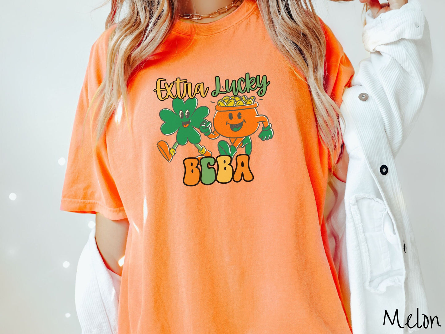 A woman wearing a vintage, melon colored shirt with the text Extra Lucky BCBA in yellow, orange, and green font. Between the text are a green clover and orange pot of gold smiling and walking together in step.