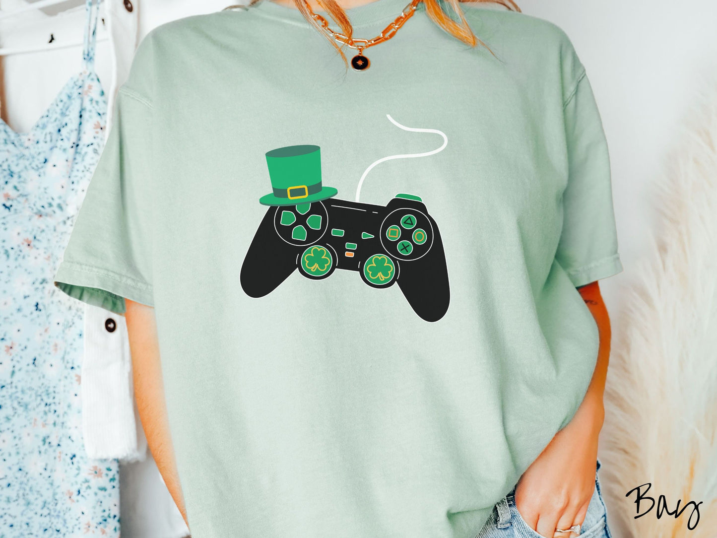 A woman wearing a vintage, bay colored shirt with a black video game controller with green buttons and joysticks, the analog control sticks are in the shape of green leaf clovers, and there is a green top hat hanging off the top left trigger.