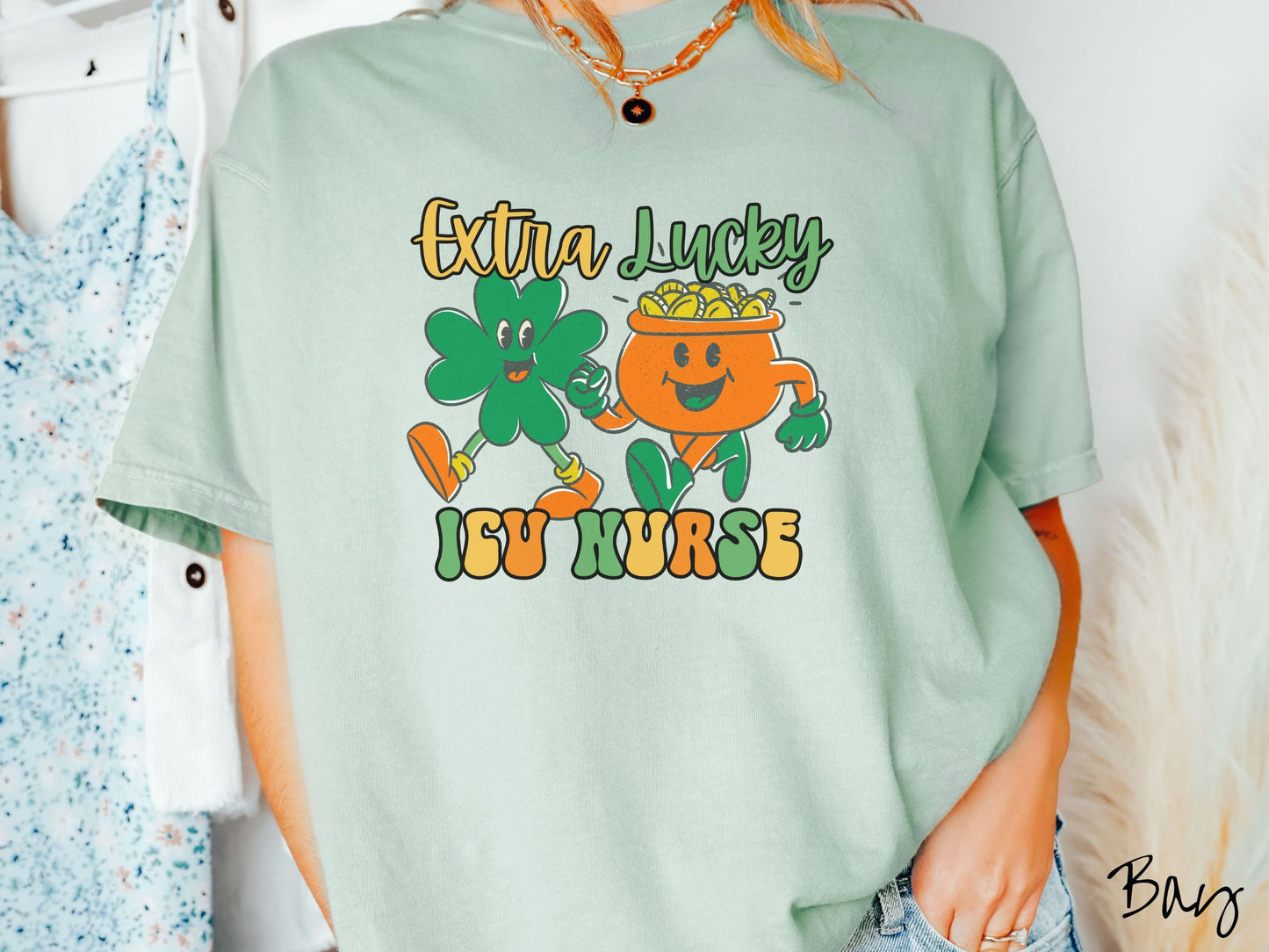 A woman wearing a vintage, bay colored shirt with the text Extra Lucky ICU Nurse in yellow, orange, and green font. Between the text are a green clover and orange pot of gold smiling and walking together in step.