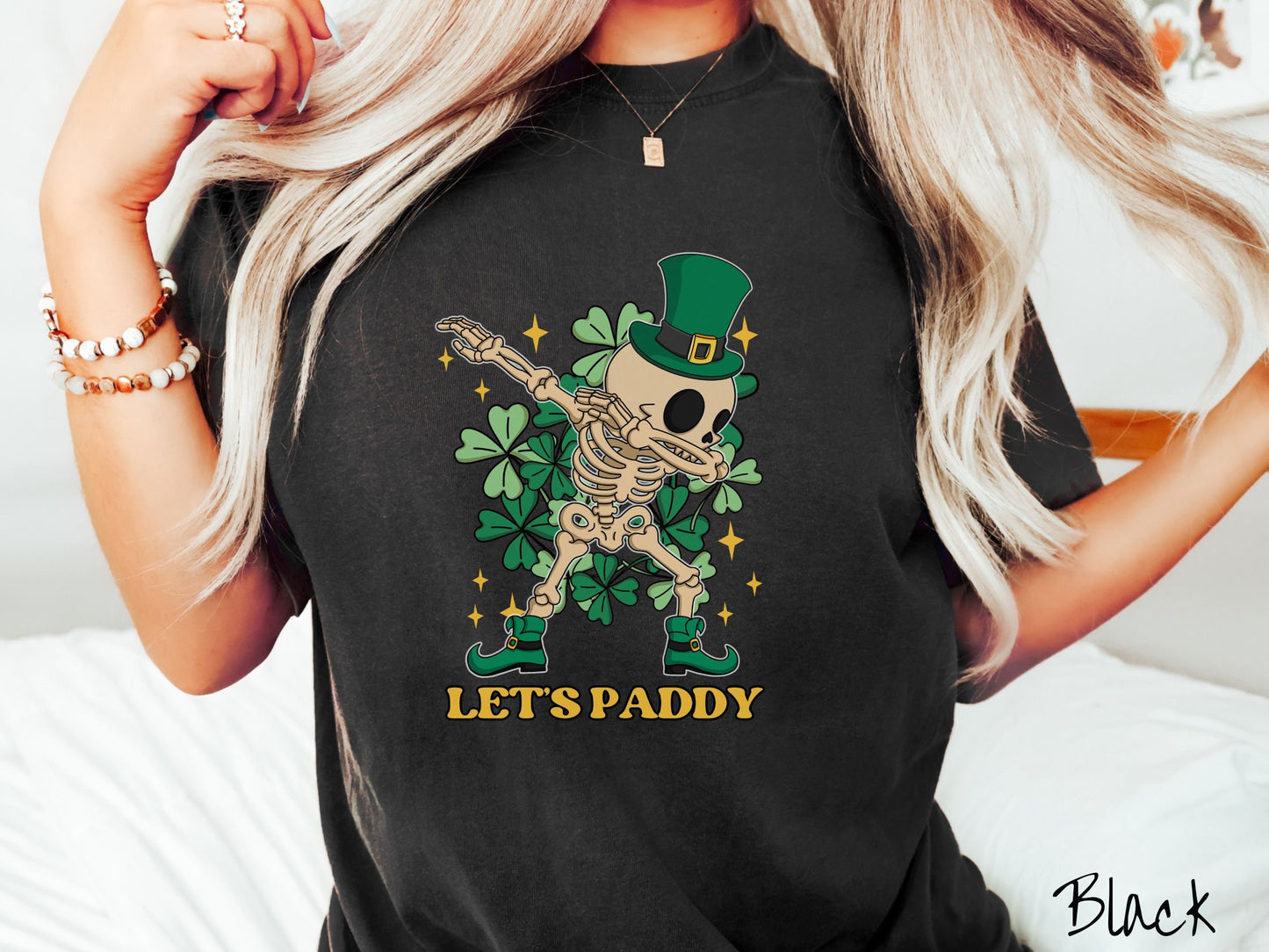 A woman wearing a vintage, black colored shirt with the text Lets Paddy and below that is a leprechaun wearing a green top hat and green elf shoes doing the dab dance and there are green clovers and golden stars in the background.