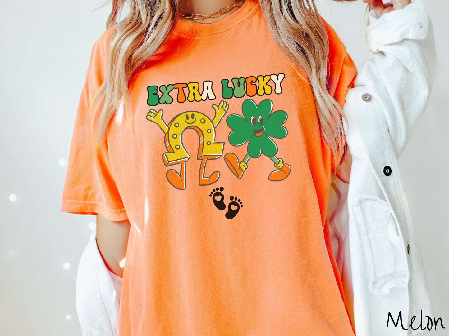 A woman wearing a vintage, melon colored shirt with text Extra Lucky in green, white, and orange font. Below are a yellow horseshoe with a smile and its hands up, a green clover smiling, and two white baby footprints with hearts.