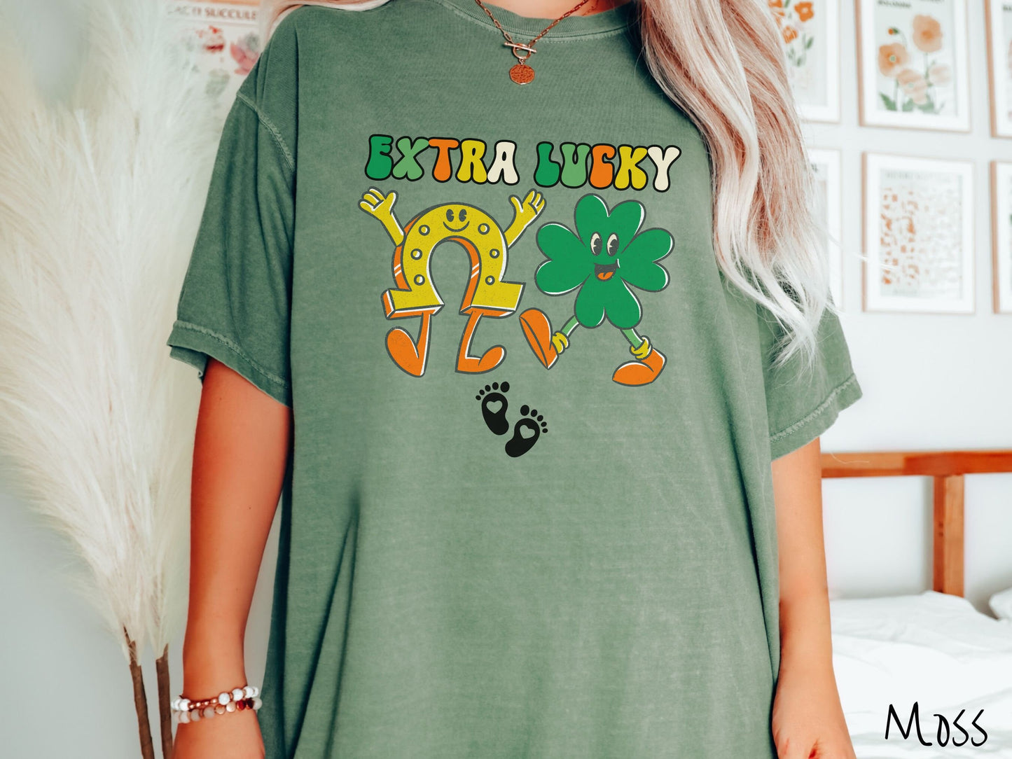 A woman wearing a vintage, moss colored shirt with text Extra Lucky in green, white, and orange font. Below are a yellow horseshoe with a smile and its hands up, a green clover smiling, and two white baby footprints with hearts.