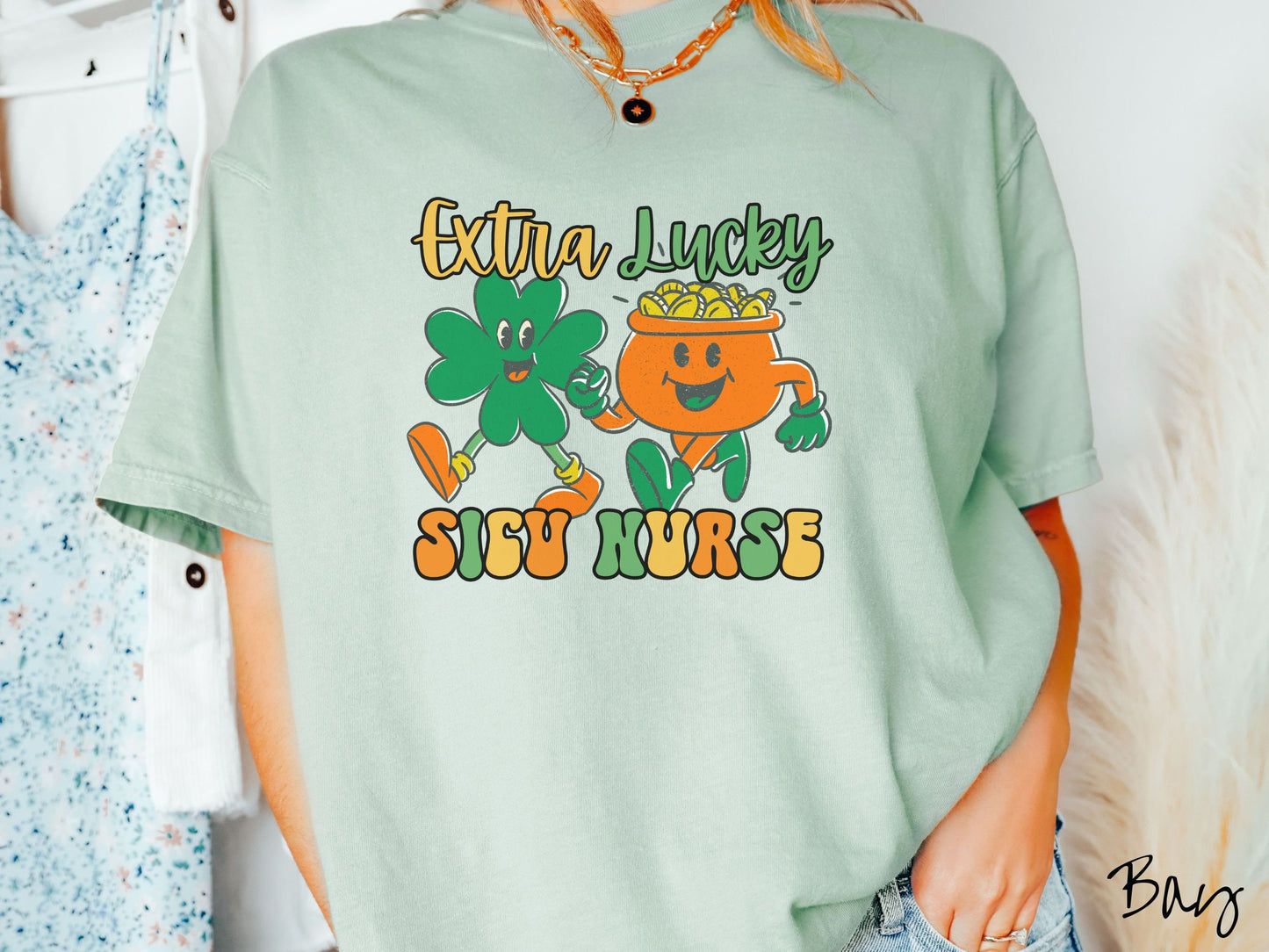 A woman wearing a vintage, bay colored shirt with the text Extra Lucky SICU Nurse in yellow, orange, and green font. Between the text are a green clover and orange pot of gold smiling and walking together in step.