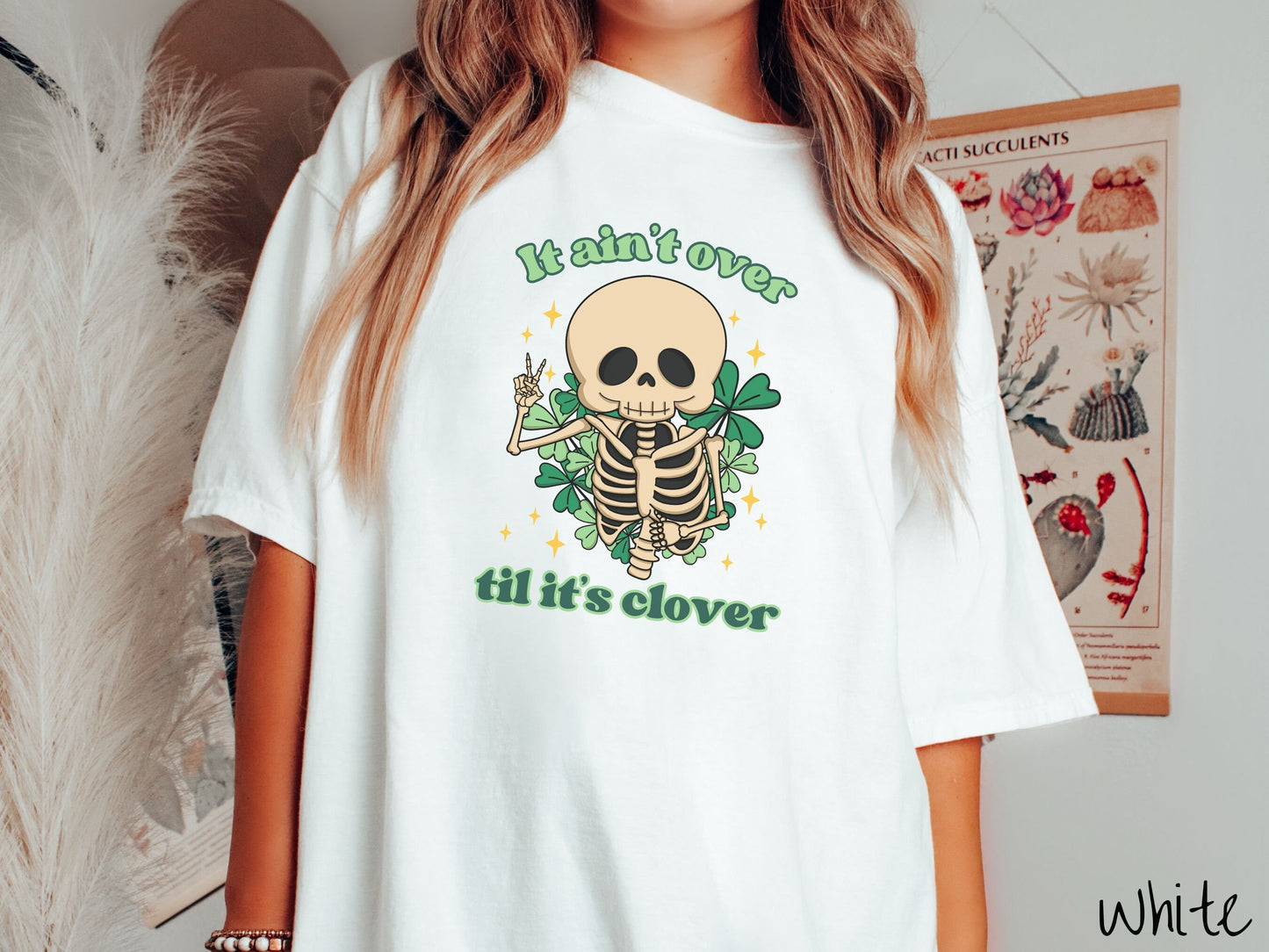 A woman wearing a vintage, white colored shirt with the text It aint over til its clover and in between the text is a smiling skeleton holding a peace sign with its right hand, with many green three leaf clovers and golden stars in the background.