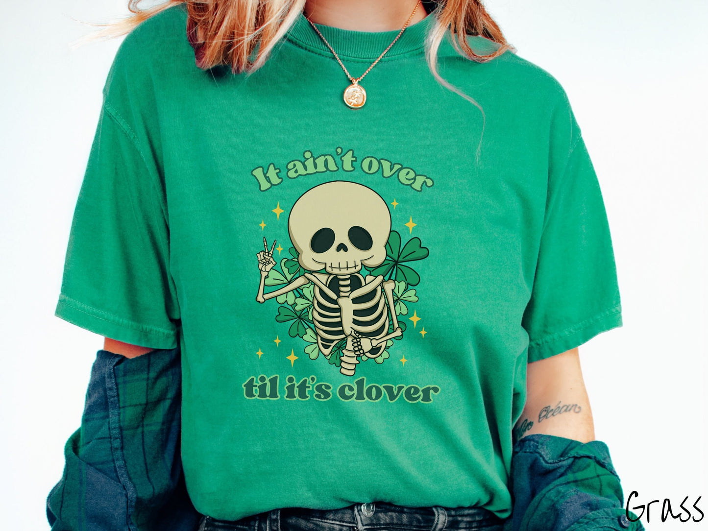 A woman wearing a vintage, grass colored shirt with the text It aint over til its clover and in between the text is a smiling skeleton holding a peace sign with its right hand, with many green three leaf clovers and golden stars in the background.