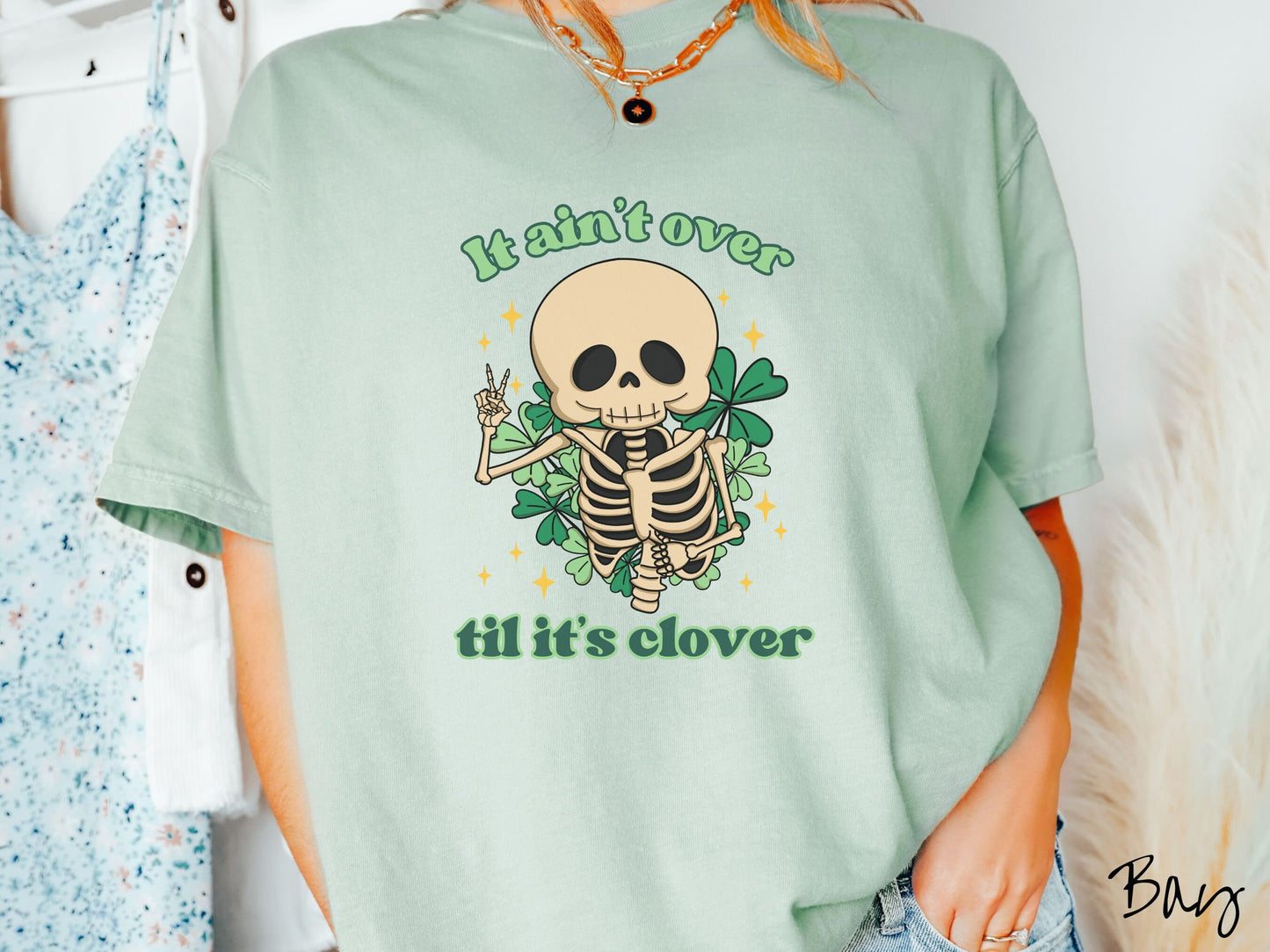 A woman wearing a vintage, bay colored shirt with the text It aint over til its clover and in between the text is a smiling skeleton holding a peace sign with its right hand, with many green three leaf clovers and golden stars in the background.
