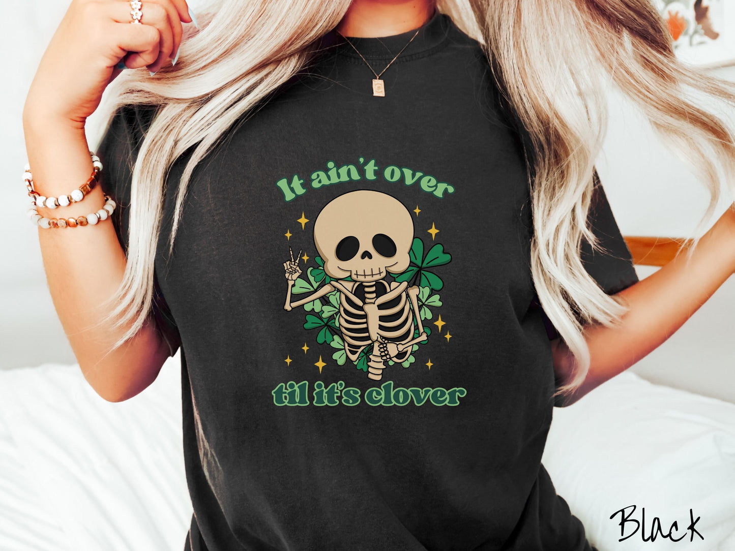 A woman wearing a vintage, black colored shirt with the text It aint over til its clover and in between the text is a smiling skeleton holding a peace sign with its right hand, with many green three leaf clovers and golden stars in the background.