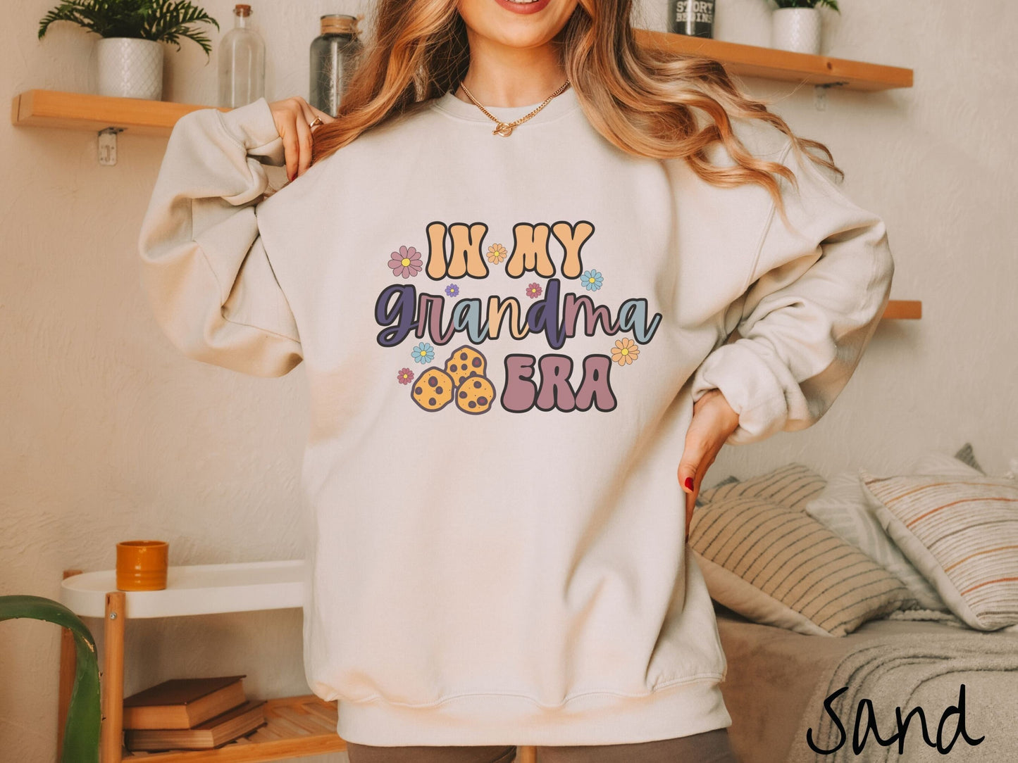 A woman wearing a cute sand colored sweatshirt with text on the front saying In My Grandma Era, with flowers and chocolate chip cookies mixed within the text.