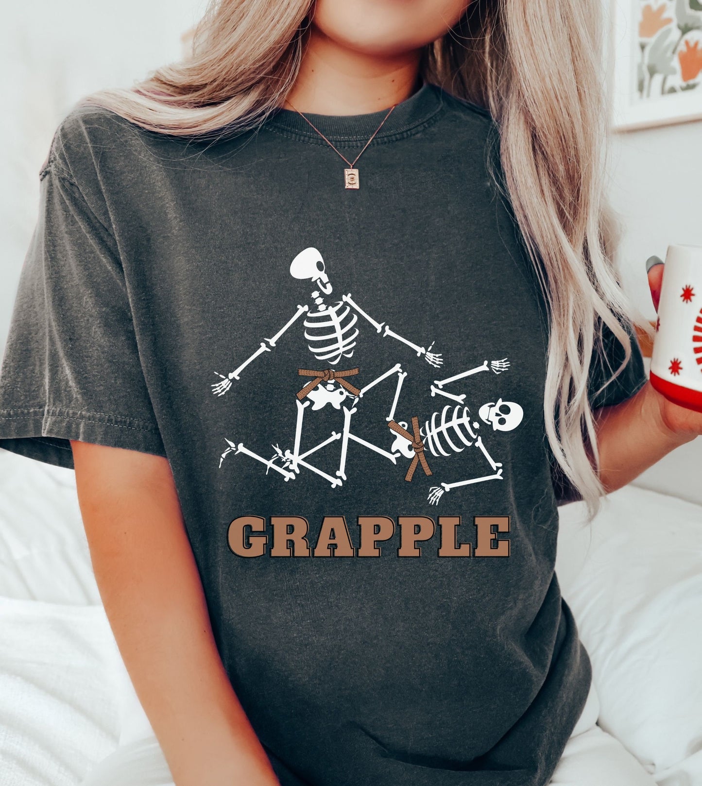 A woman wearing a cute pepper colored shirt with brown text Grapple and two skeletons wearing brown belts grappling on the ground.